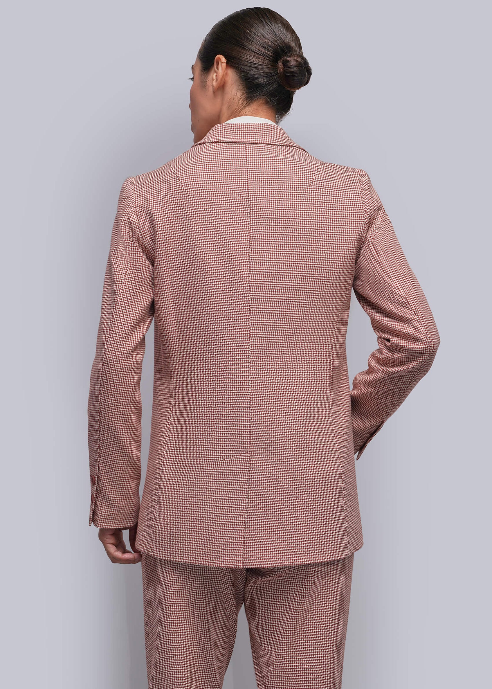 S=A person viewed from the back wearing a matching dusty red and white micro-check patterned suit, consisting of a blazer and trousers, against a solid light gray background.