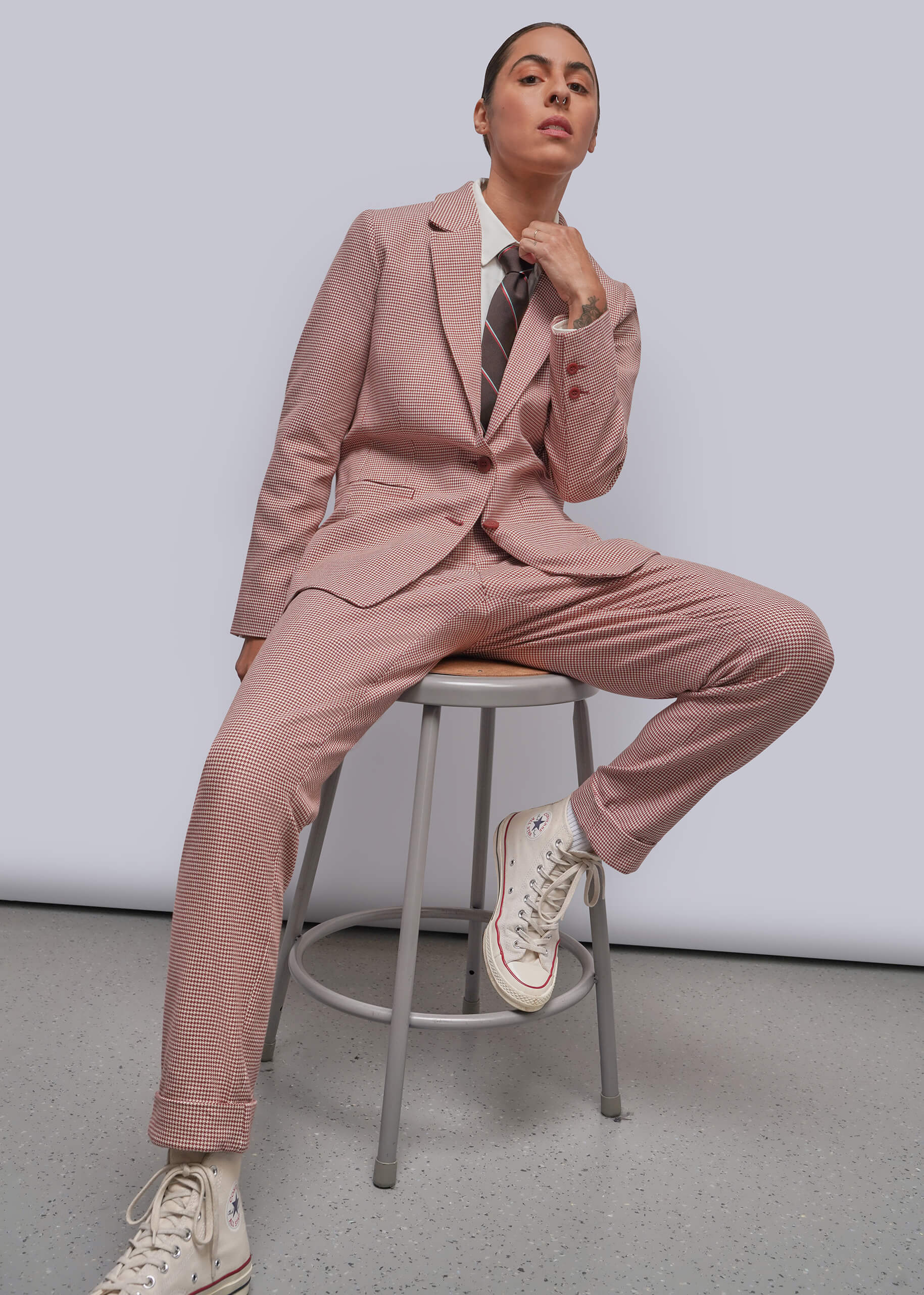 S=A model wearing a dusty red tailored suit with matching trousers, a white shirt, a striped tie, and white sneakers, sitting on a metal stool against a light gray studio background.