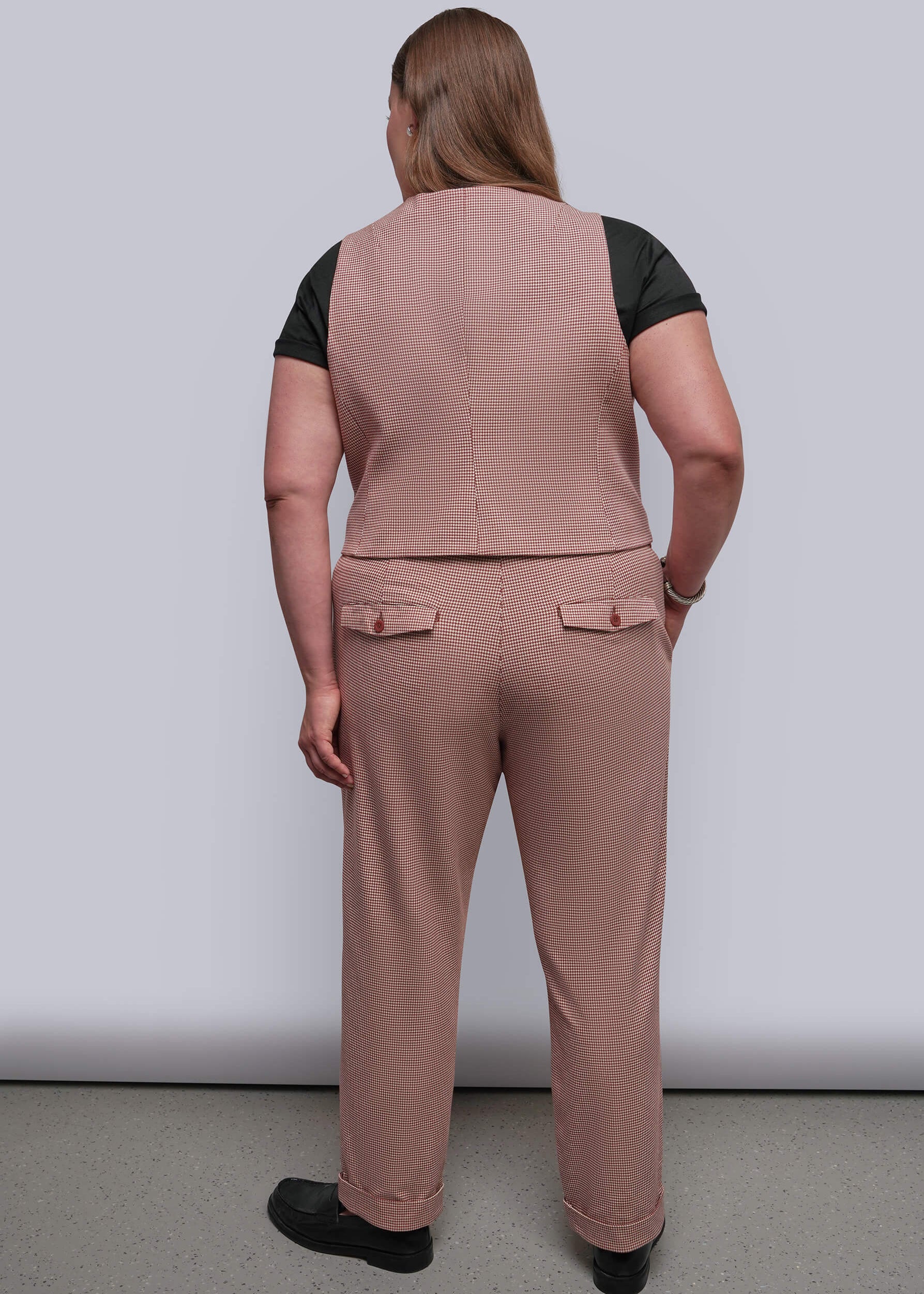 1X=A woman viewed from the back wearing a dusty red vest and matching trousers over a black short-sleeve top, standing against a plain light gray studio background.