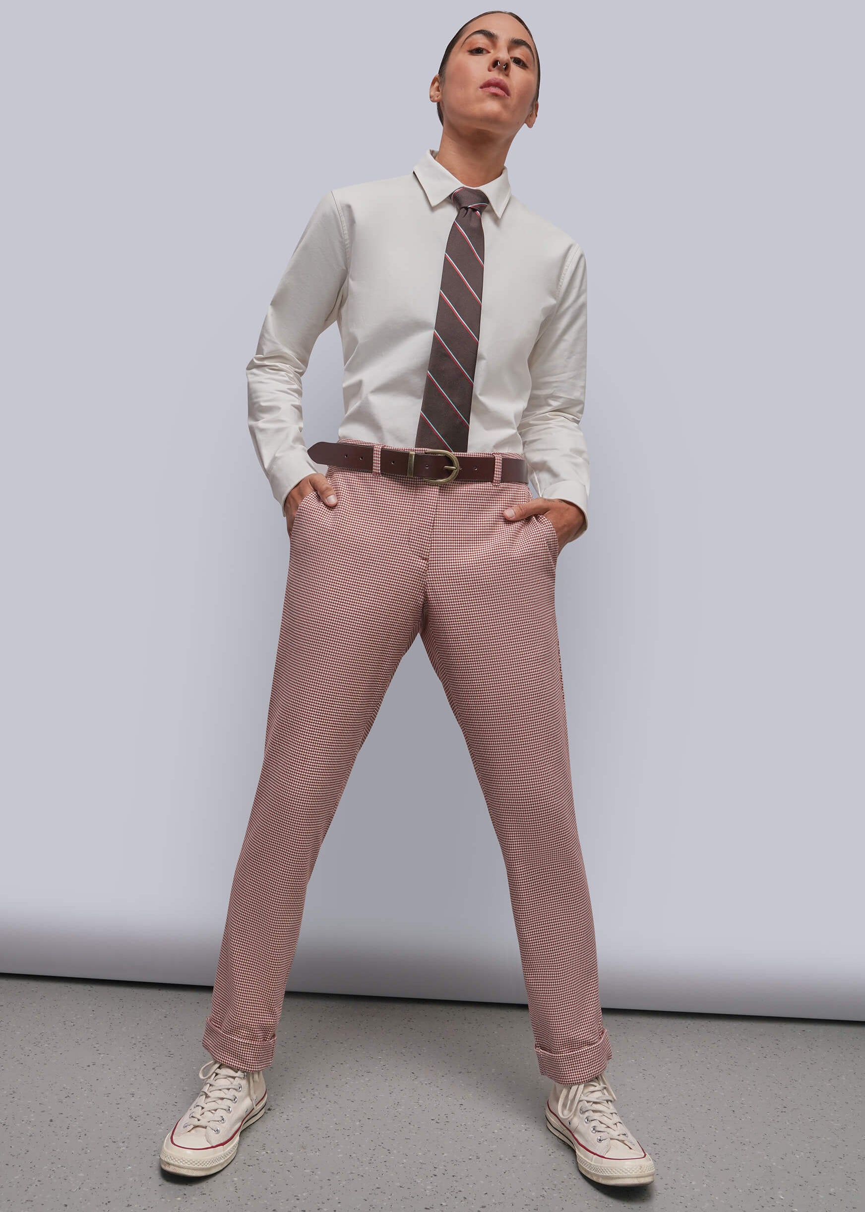 S=A model wearing a white dress shirt, a brown striped tie, dusty red houndstooth trousers, a brown belt, and white sneakers, standing against a light gray studio background.