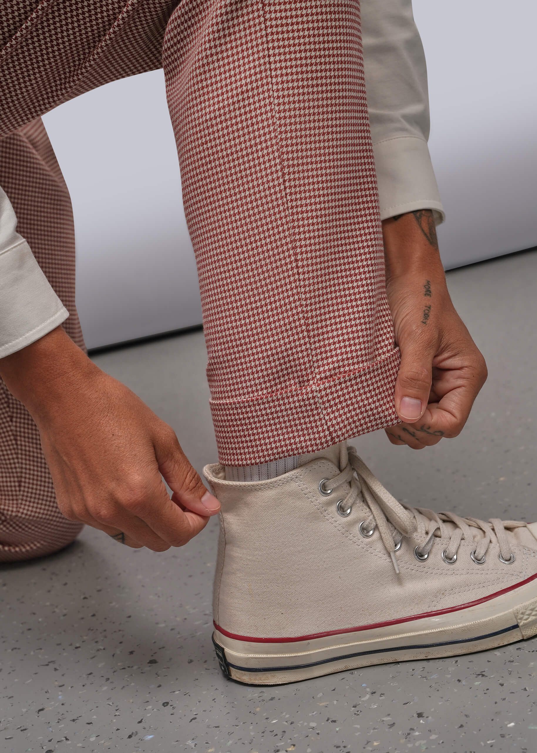S=A close-up of a person's hands adjusting the cuff of their houndstooth patterned trouser while wearing a light beige shirt and white sneakers.