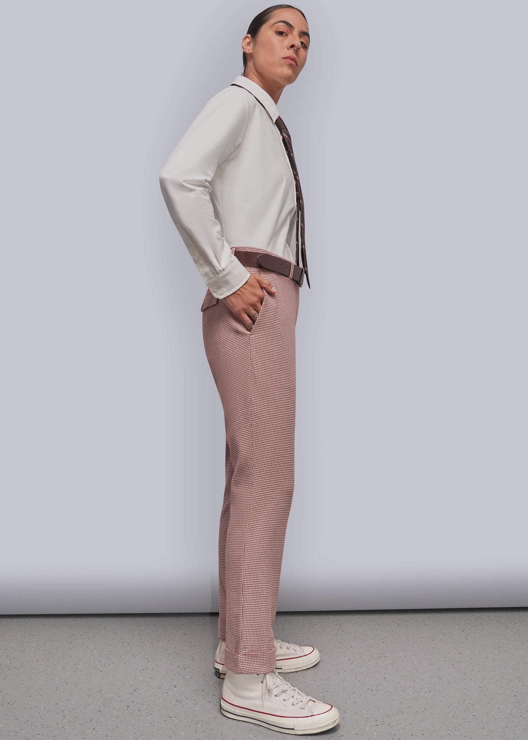 S=A model is shown in profile wearing a whitw long-sleeve button-down shirt, a dark brown patterned tie, dusty red patterned trousers, a brown belt, and off-white canvas sneakers against a light gray studio background.