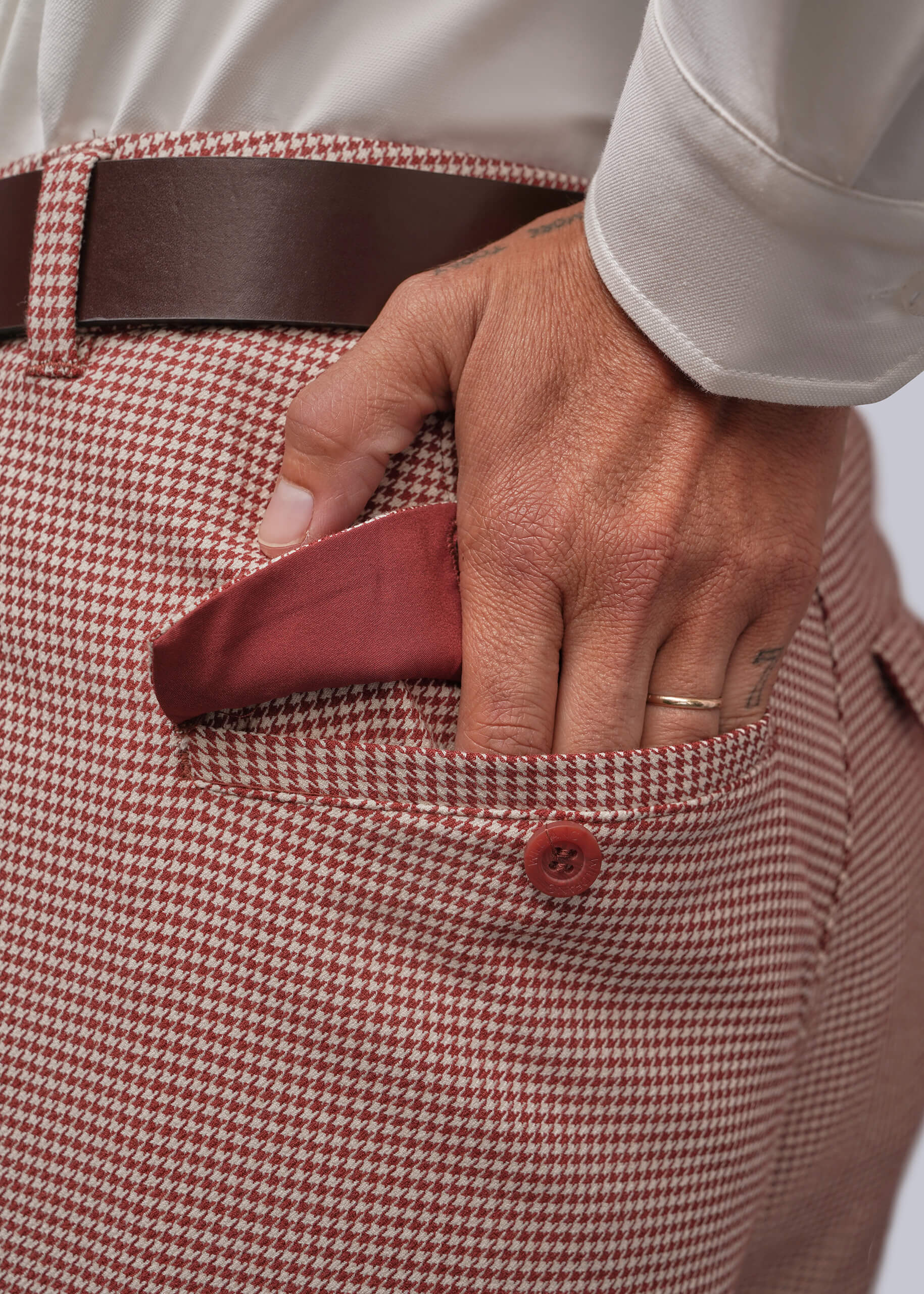 S=Close-up of a person wearing a red and white houndstooth patterned trouser, with a brown leather belt and a red pocket lining visible as a hand reaches into the back pocket.
