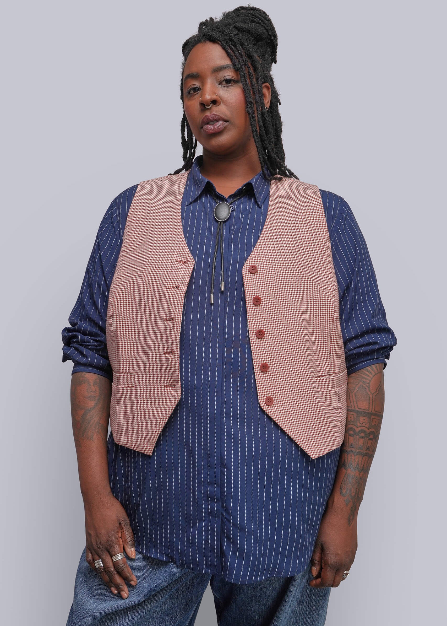1X=A model wearing a dusty red houndstooth vest over a blue and white pinstriped button-down shirt, paired with blue jeans, against a solid light gray background.