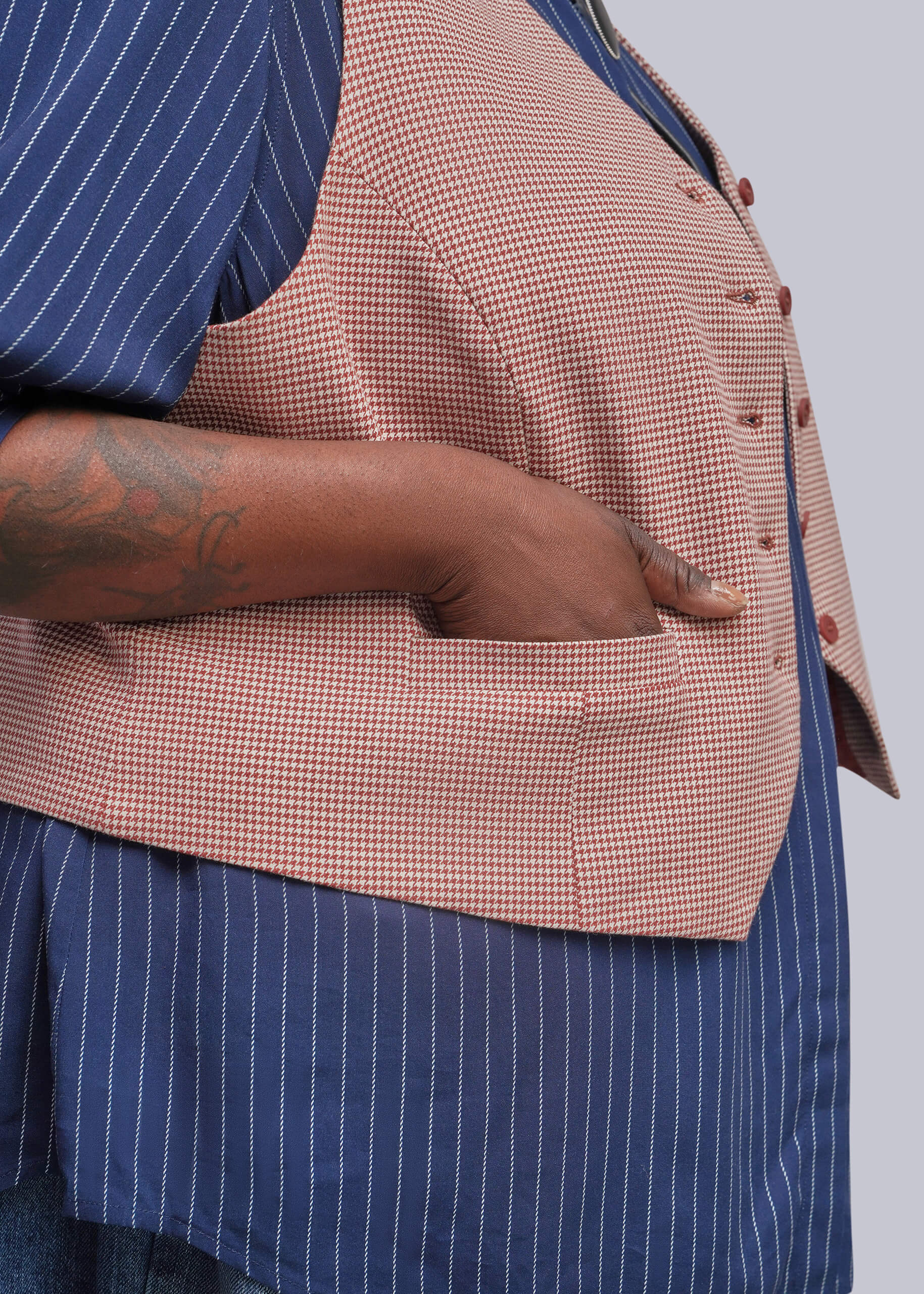 1X=A close-up of a person wearing a red and white houndstooth vest over a blue and white pinstriped shirt, with one hand tucked into a pocket.