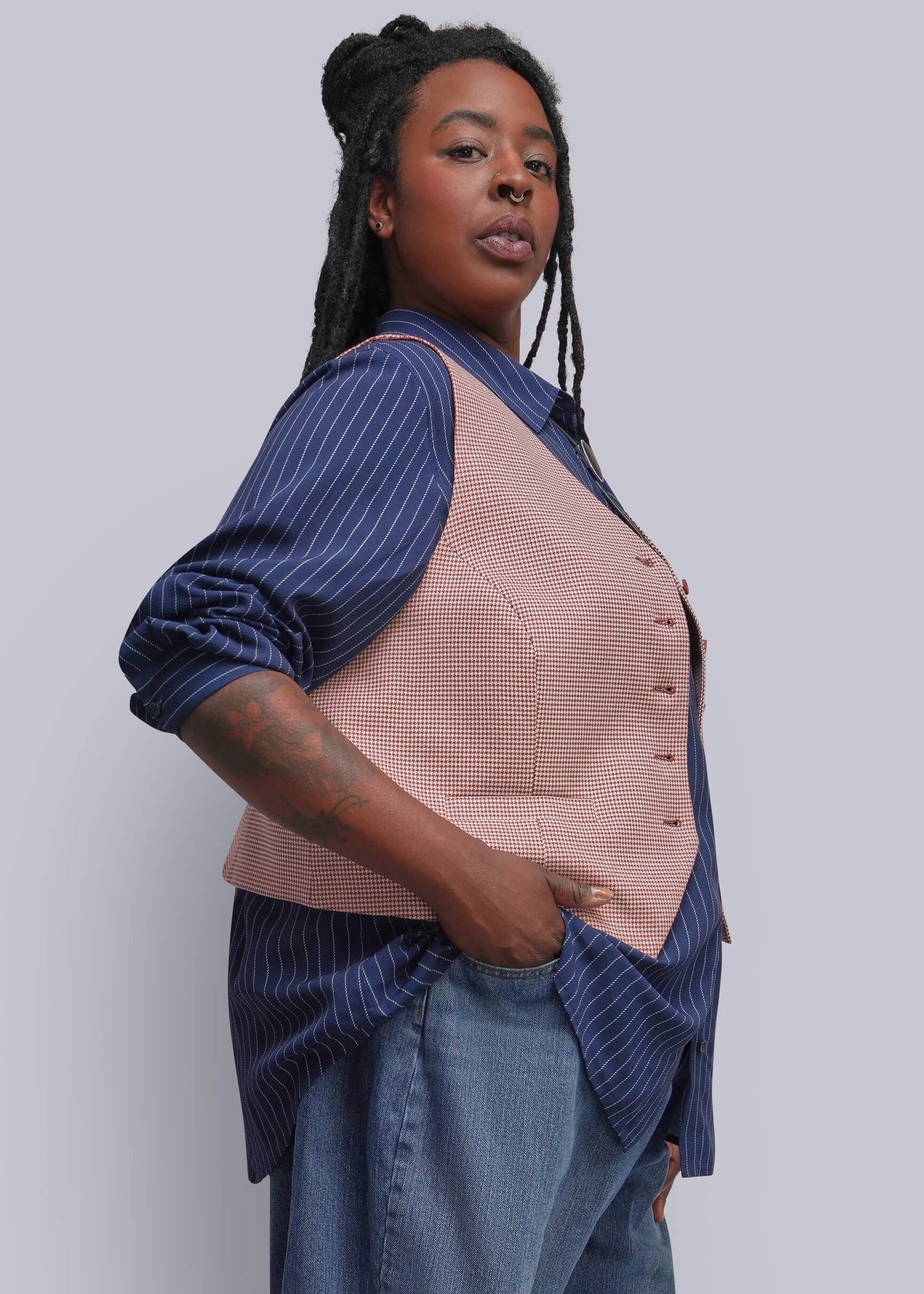 1X=A woman wearing a dusty red textured vest over a navy blue and white pinstriped shirt, paired with blue jeans, photographed against a solid light gray background.