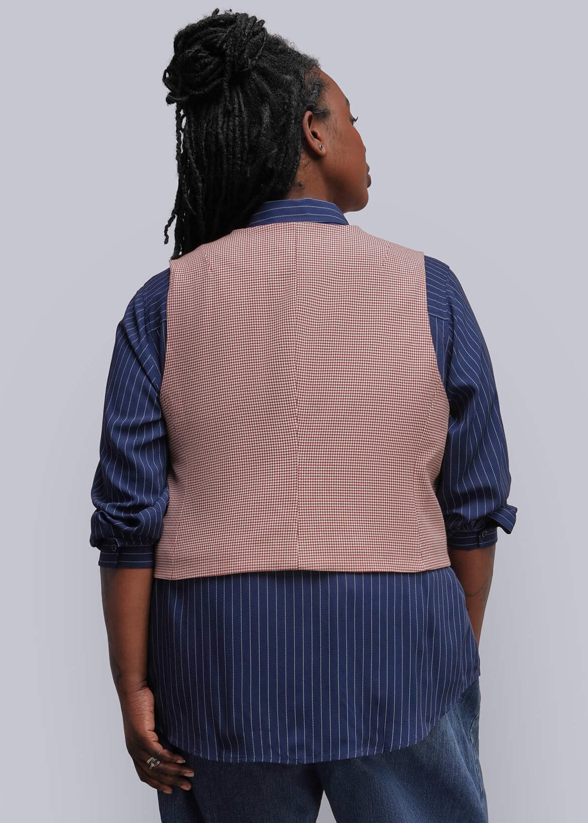 1X=A model is shown from the back wearing a dusty red vest over a navy blue and white striped shirt, paired with blue jeans and black heels, against a plain light gray studio background.