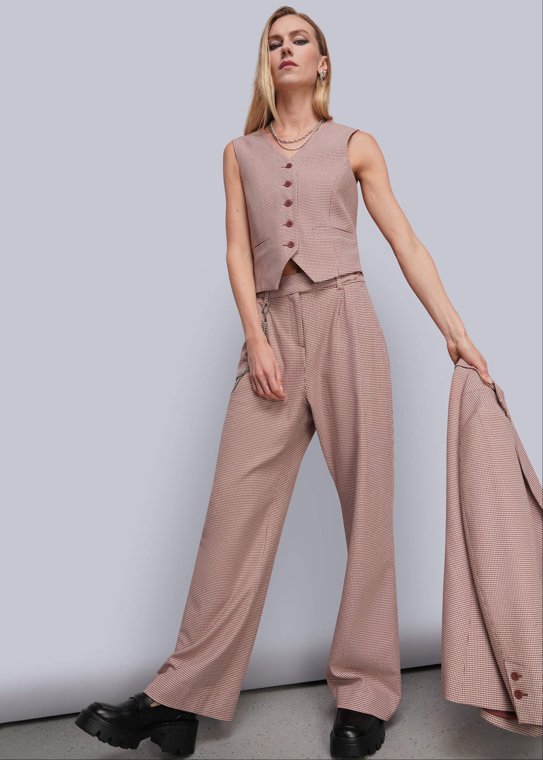 A woman models a sleeveless dusty red vest and matching wide-leg trousers, holding a matching blazer against a plain light gray studio background.