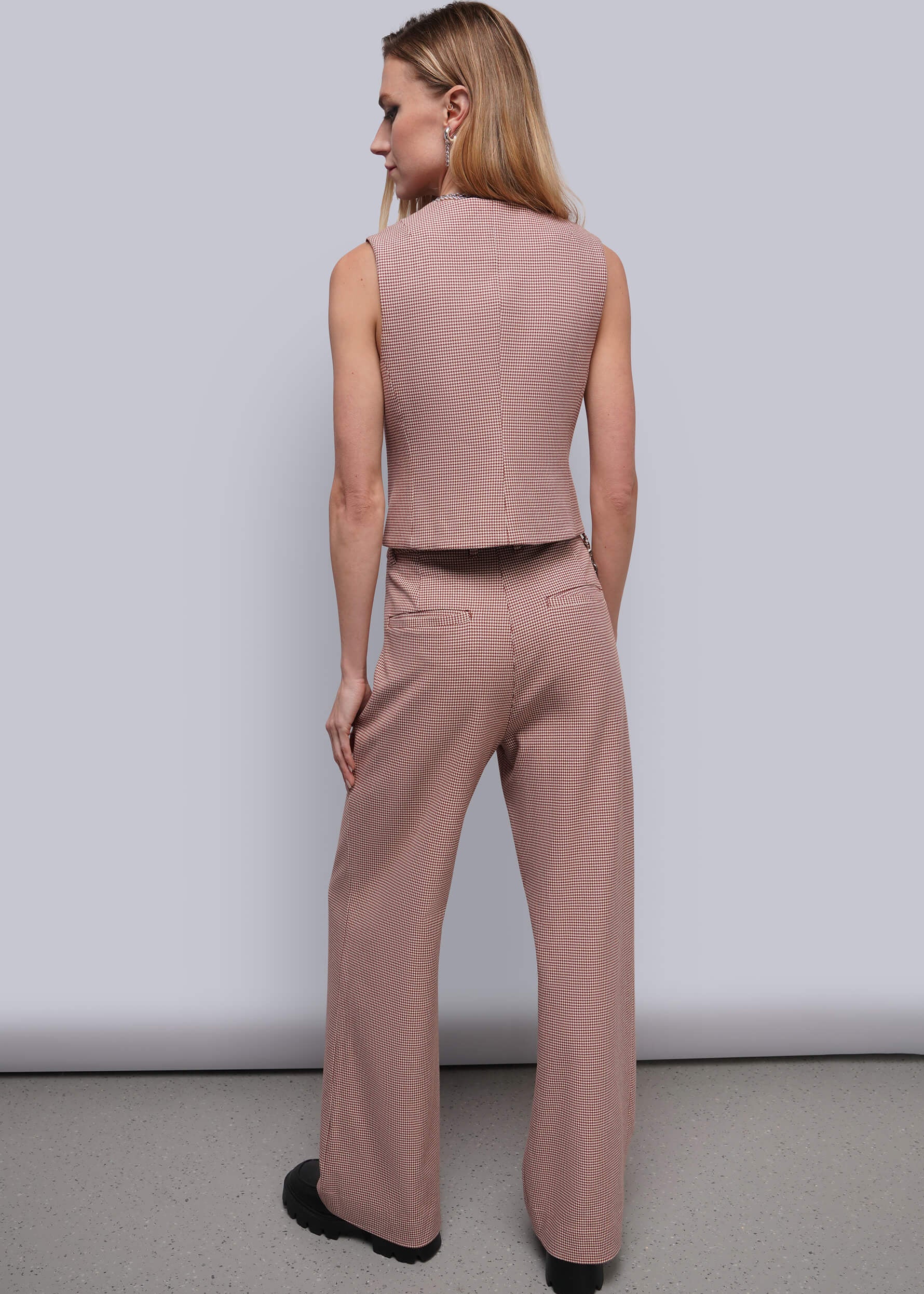 S=A woman shown from the back wearing a sleeveless dusty red houndstooth vest and matching wide-leg trousers, standing against a plain light gray studio wall.