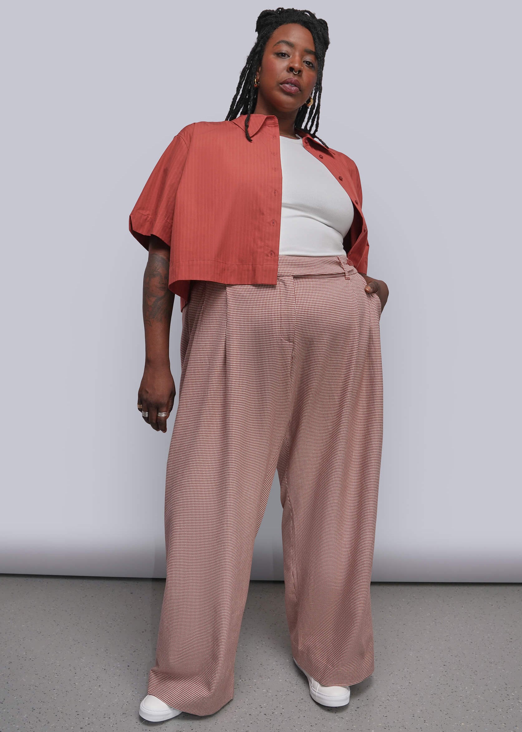 1X=A model stands against a light gray studio wall wearing a terracotta cropped button-up shirt layered over a white top and dusty red wide-leg trousers, paired with white sneakers.