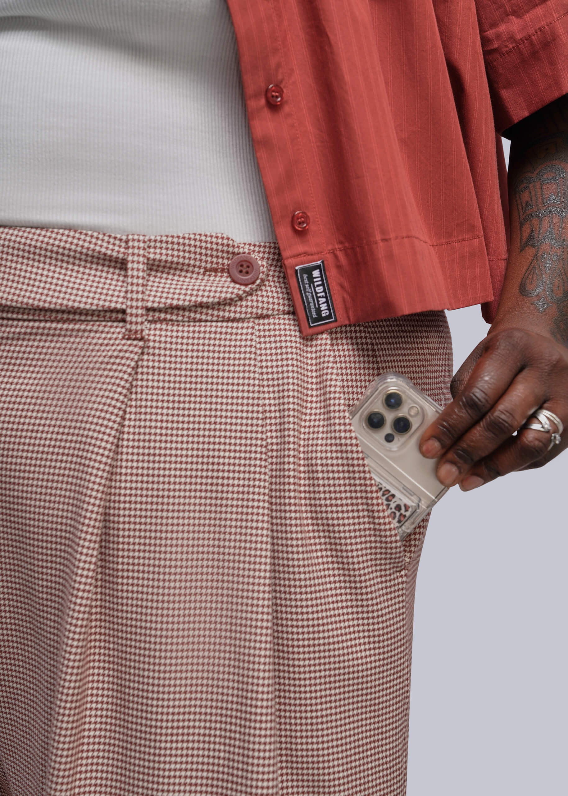 1X=Close-up of a person wearing red and beige houndstooth pleated trousers with a side pocket holding a phone, paired with a white shirt and a rust-colored short-sleeve button-up shirt. A black brand label on the shirt reads 'WILDFANG'.