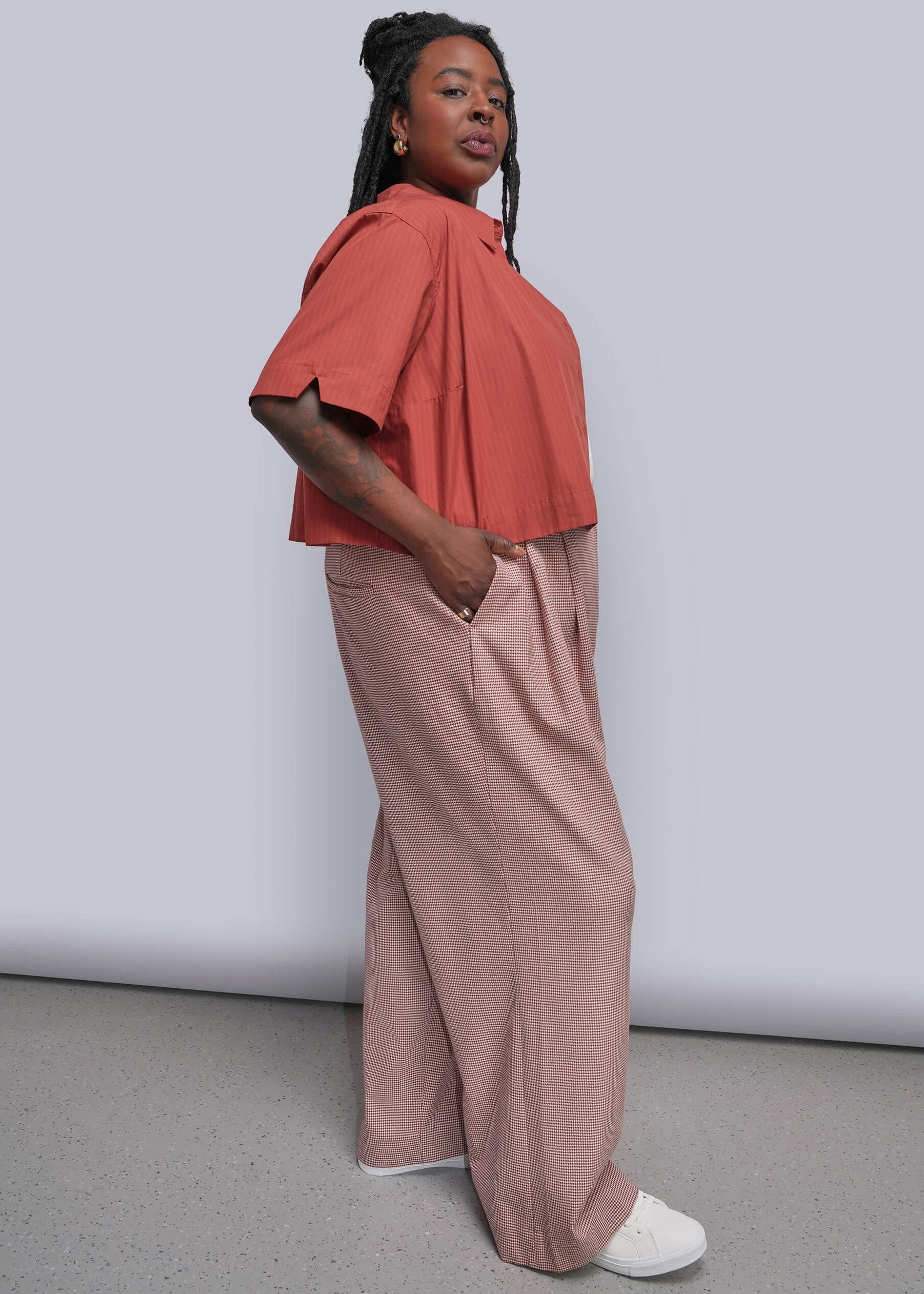 1X=A woman modeling a two-piece outfit consisting of a coral orange short-sleeve top and dusty red houndstooth wide-leg trousers, paired with white sneakers, against a plain light gray studio background.