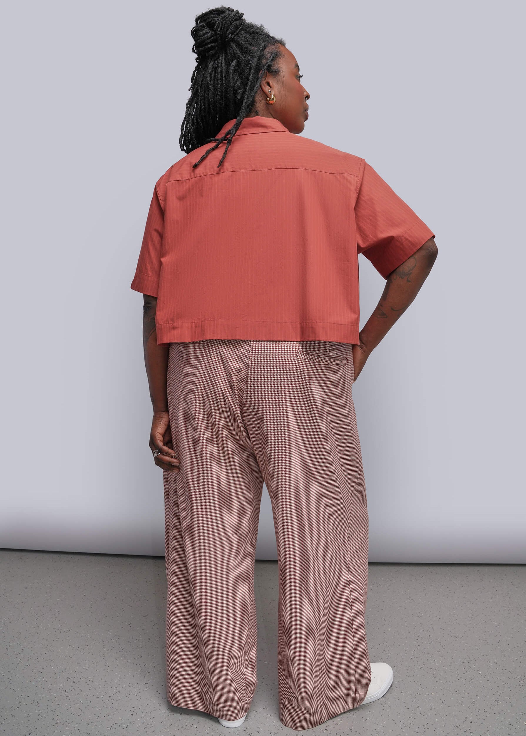 1X=A model shown from the back wearing a coral short-sleeve cropped button-up shirt and dusty red wide-leg trousers, paired with white sneakers, against a light gray studio background.