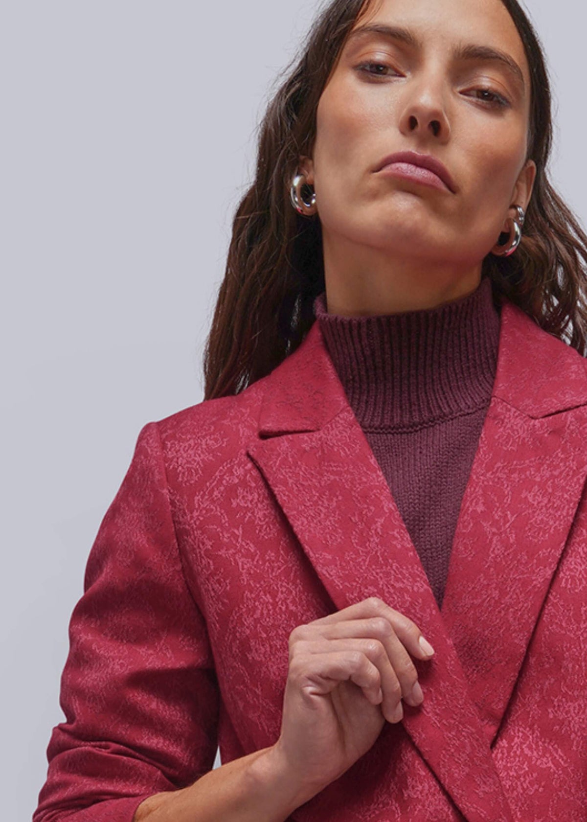 S=A close-up of a woman wearing a textured, deep pinkish-red blazer over a dark burgundy turtleneck sweater, set against a plain light gray studio background.