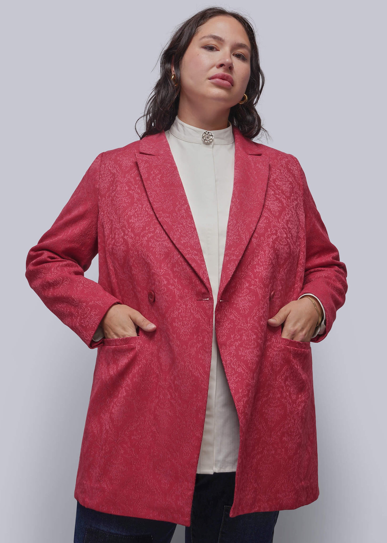 1X=A person wearing a textured, deep pinkish-red blazer over a cream mock neck top and dark jeans, standing against a solid light gray studio background.