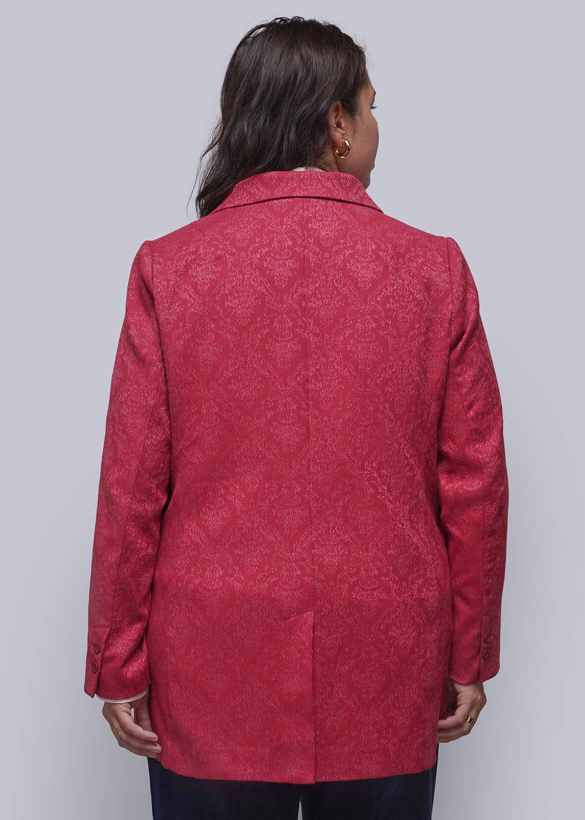 1X=A person viewed from behind wearing a textured red blazer with a subtle damask pattern, paired with dark navy trousers and black heels, standing against a plain light gray wall.