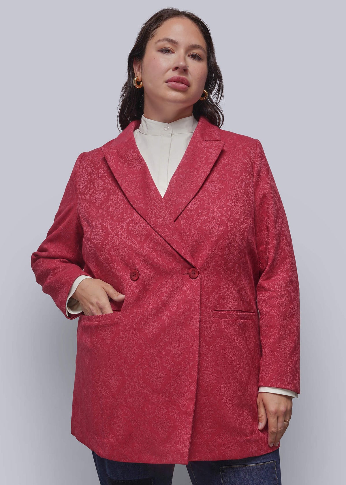 1X=A person modeling a textured, double-breasted blazer in a hot pink color, worn over a cream mock neck top and blue jeans, against a solid light gray background.