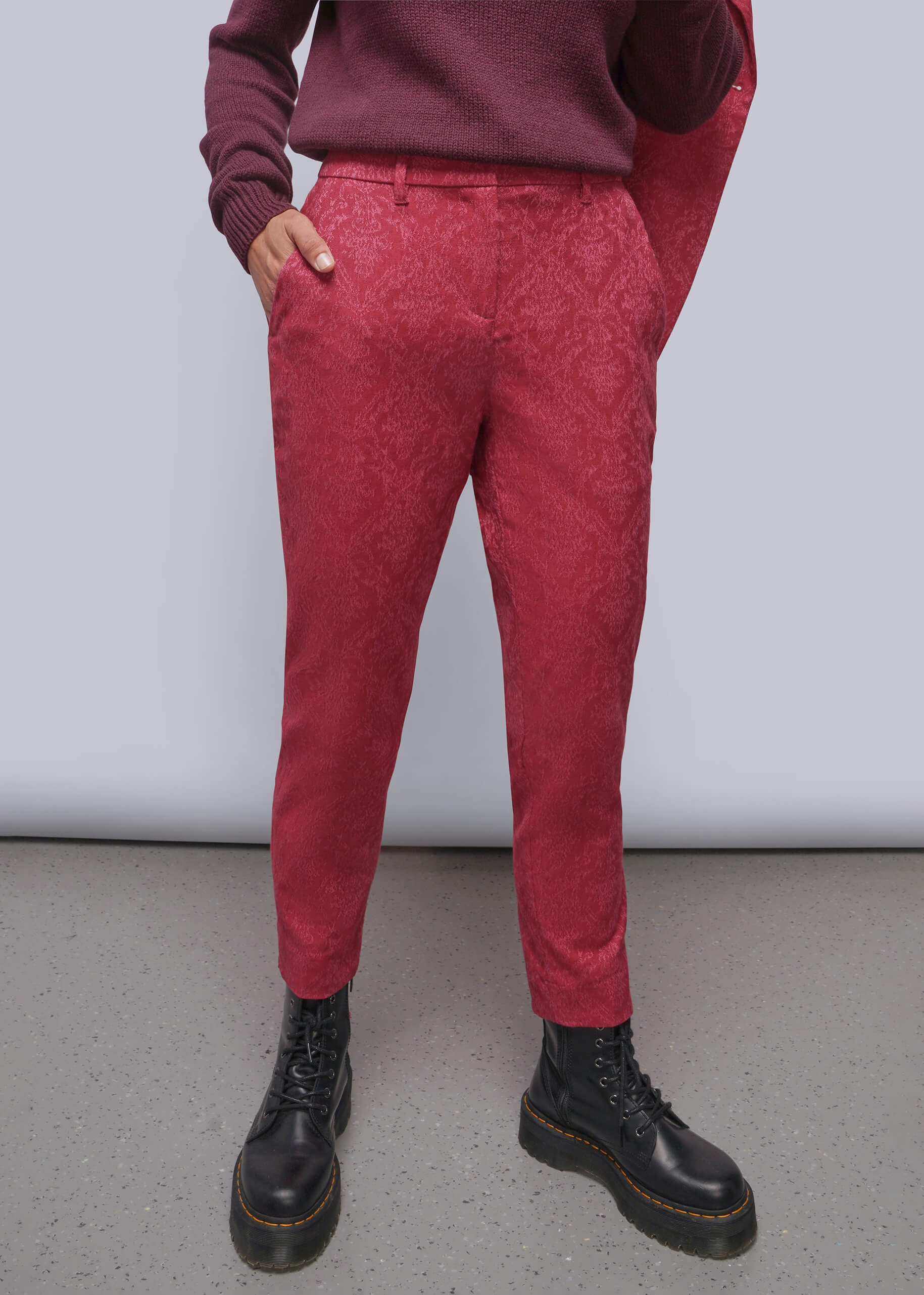 A person wearing textured red trousers, a burgundy sweater, and black leather boots, standing against a light gray studio background.