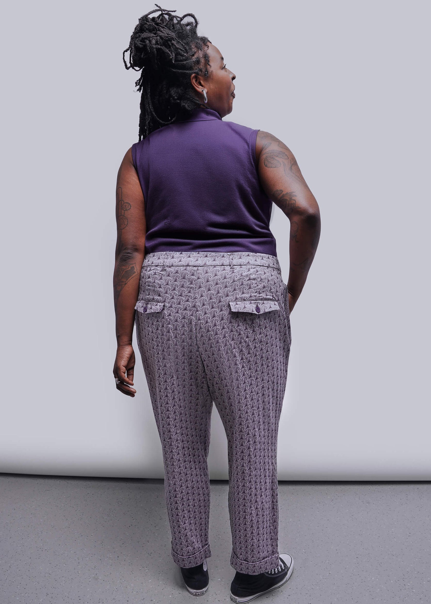 1X=Person wearing a purple sleeveless top and patterned pants against a plain background