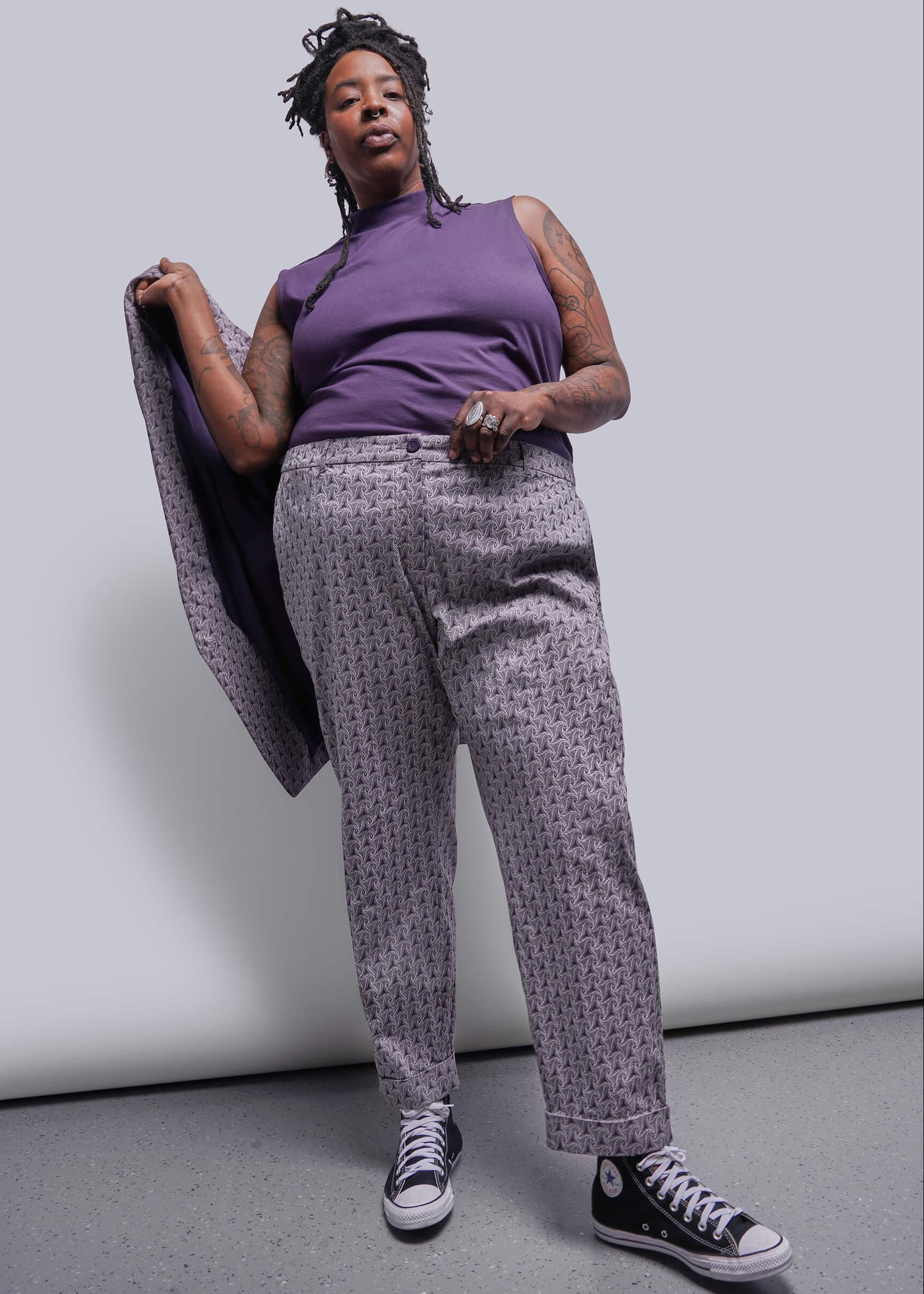 1X=Person wearing a purple top and patterned pants, holding a coat, against a plain background
