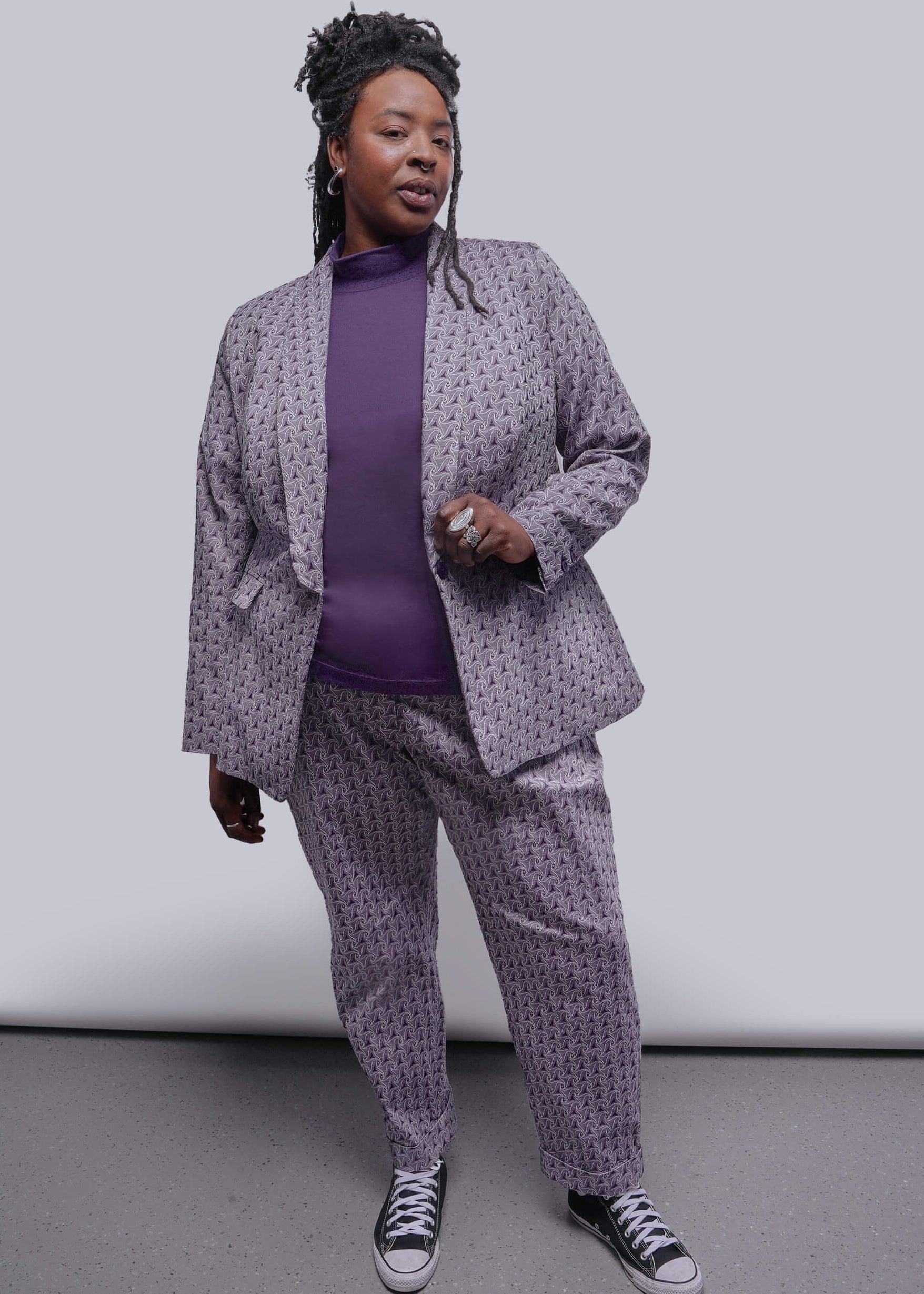 1X=Person wearing a gray patterned suit with a purple shirt and black sneakers on a plain background