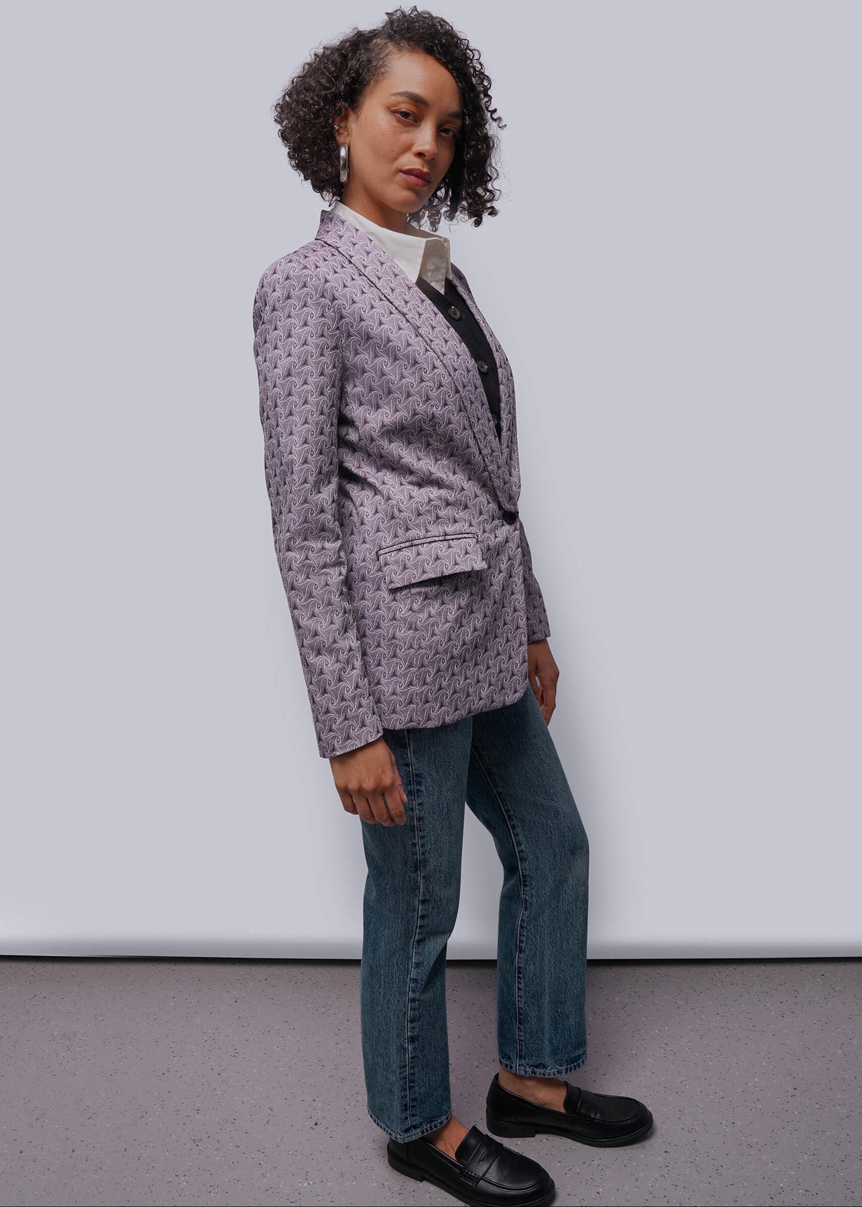 S=Person wearing a patterned blazer, black shirt, jeans, and black shoes against a white background