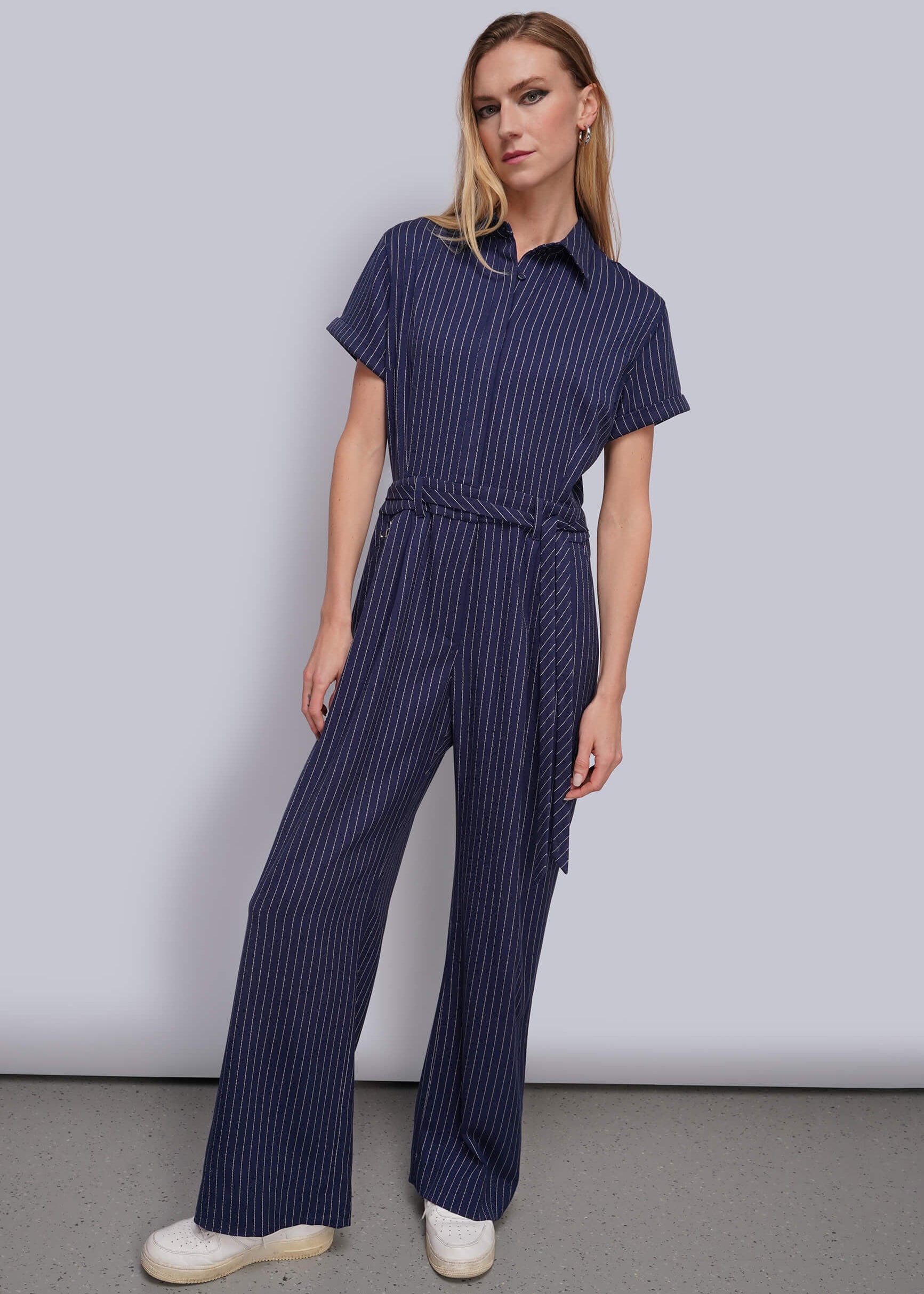 S=A woman modeling a navy blue and white vertically striped short-sleeve jumpsuit with a matching tie belt, paired with white sneakers, against a plain light gray studio background.