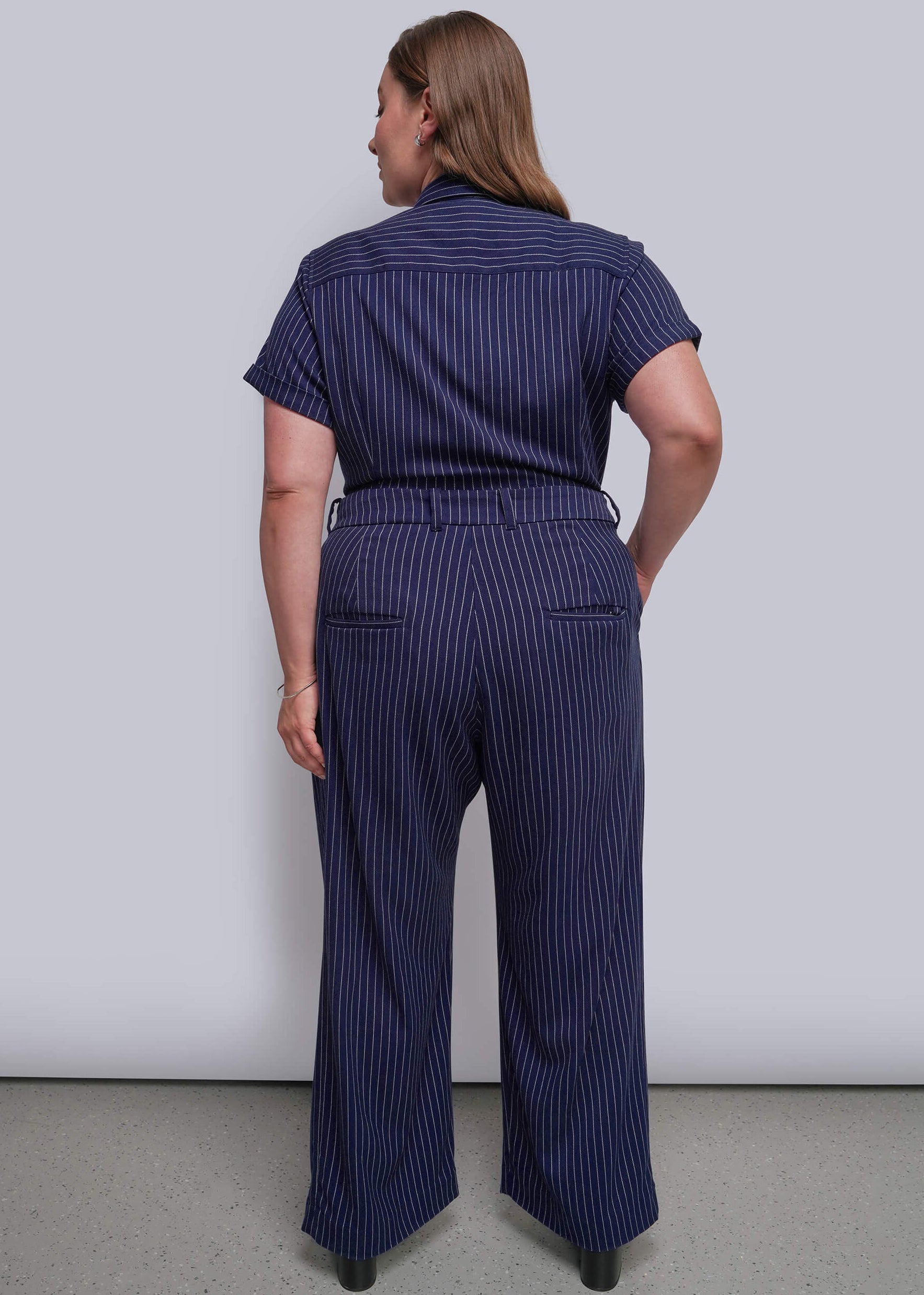 1X=A woman viewed from the back wearing a navy blue short-sleeve jumpsuit, standing against a plain light gray wall.