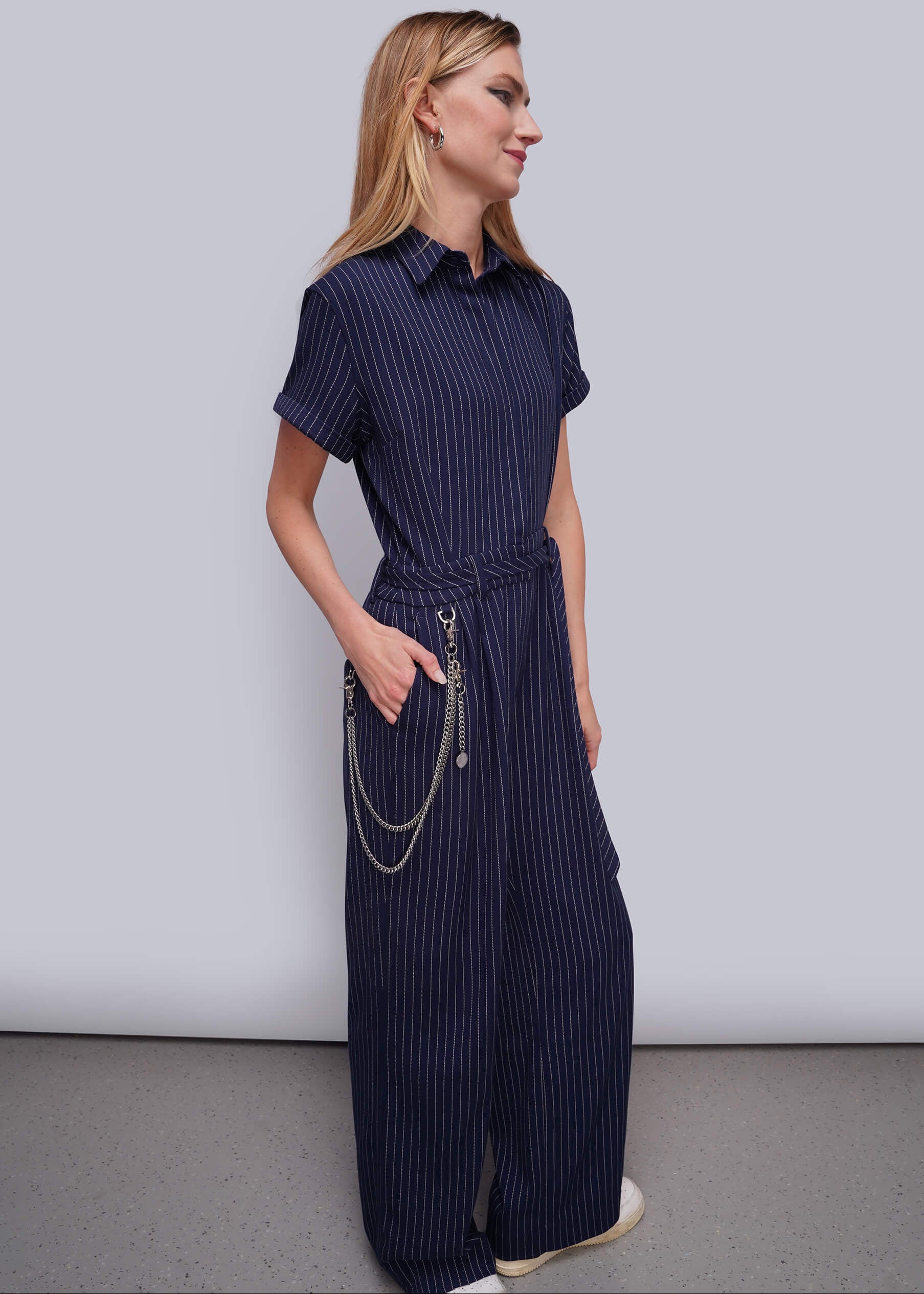 S=A woman modeling a navy blue pinstriped short-sleeve jumpsuit with a tie-waist belt and silver chain detail, standing against a light gray studio background.