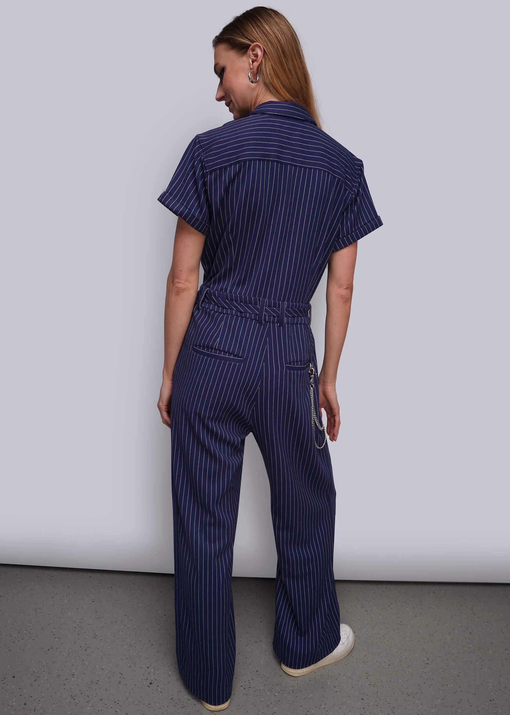 S=A woman viewed from behind wearing a navy blue short-sleeve jumpsuit with thin white vertical pinstripes, featuring a button-down back collar, a drawstring waist, and wide-leg trousers.