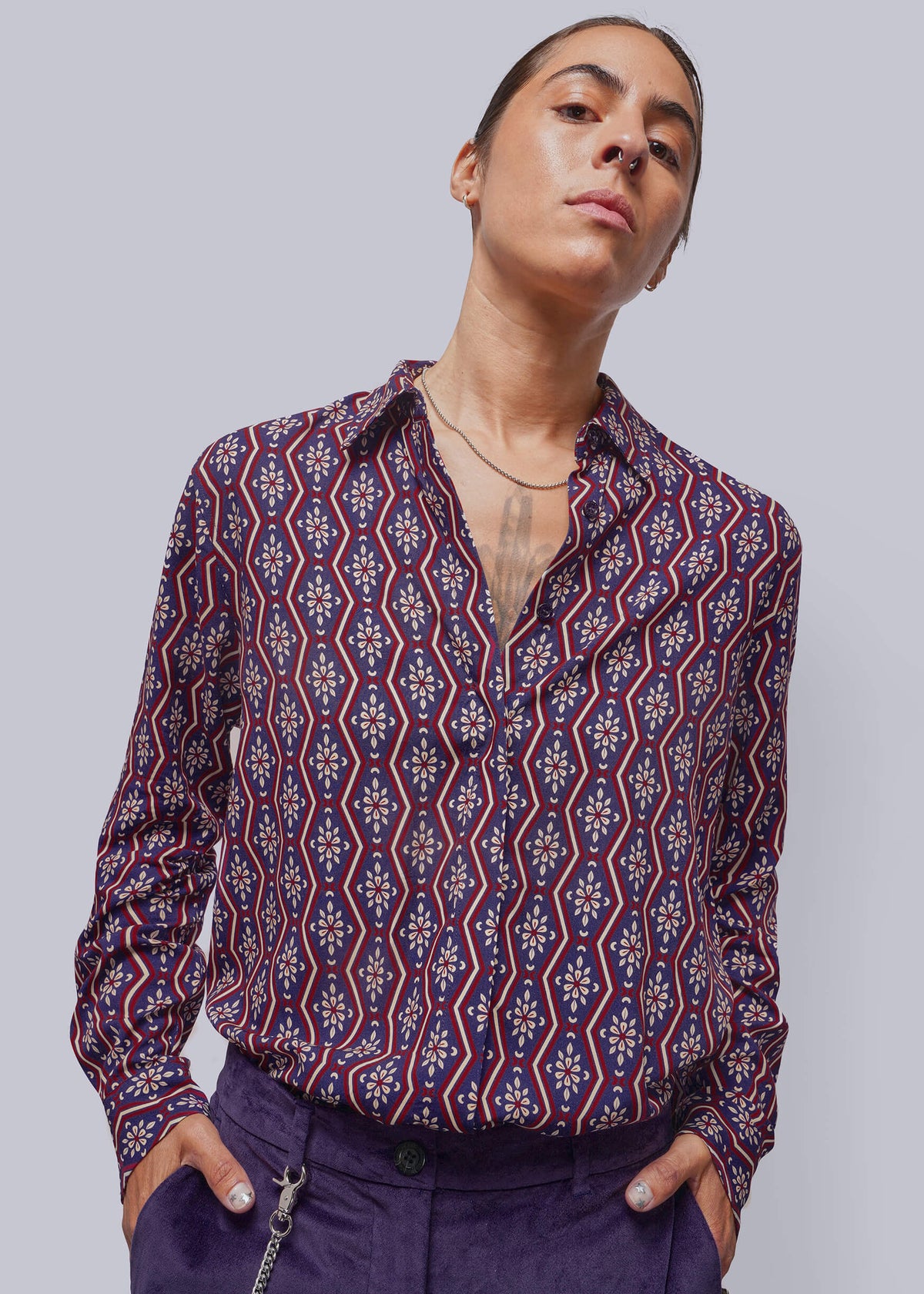 S=A model wearing a long-sleeve button-up shirt with a red, blue, and white geometric pattern, paired with purple trousers, against a solid gray background.
