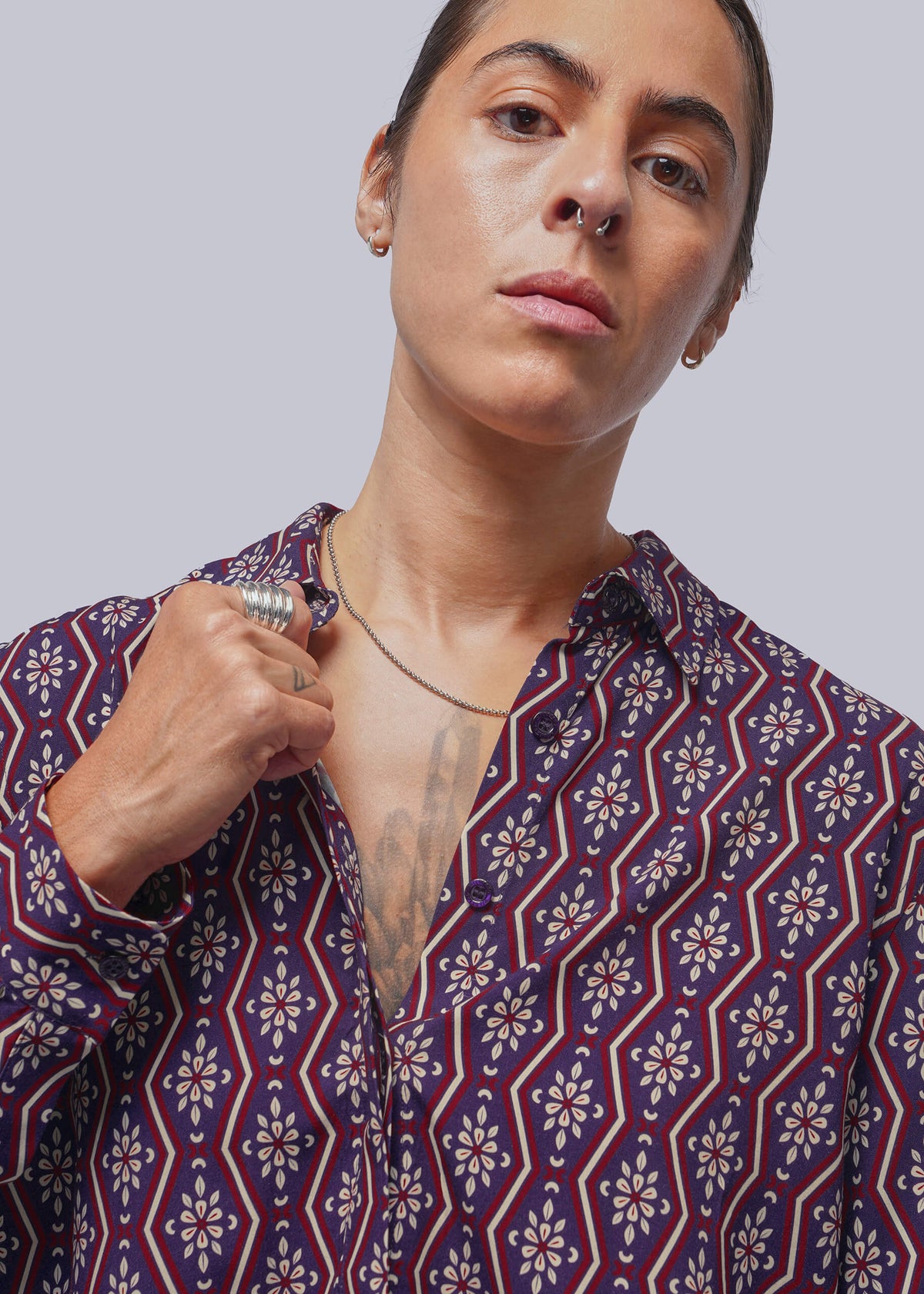 S=A close-up of a person wearing a patterned button-up shirt with a geometric and floral design in navy blue, red, and white, set against a solid light gray background.