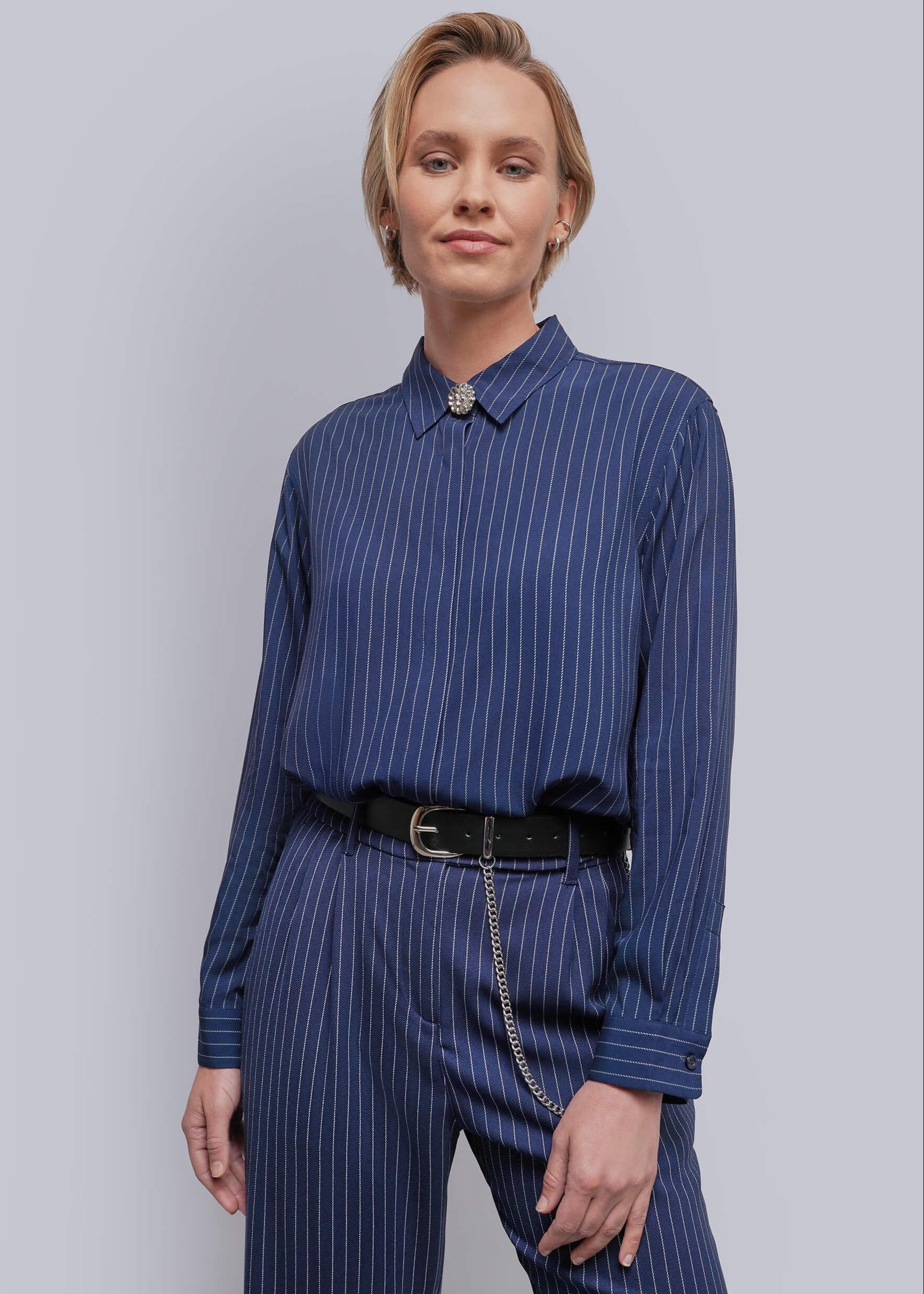 S=A woman modeling a navy blue long-sleeve button-up shirt with thin white pinstripes, paired with matching navy pinstripe trousers, a black belt, and a silver chain detail, against a solid light gray background.
