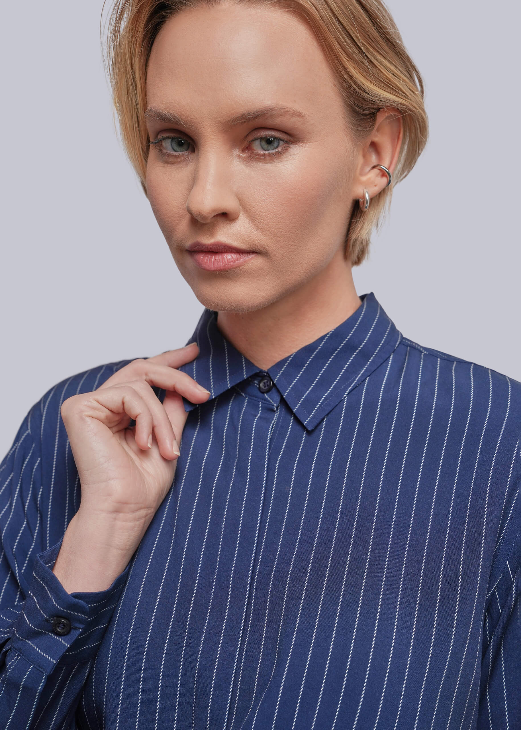 A close-up of a woman wearing a blue button-down shirt with thin white vertical pinstripes, set against a solid light gray background.