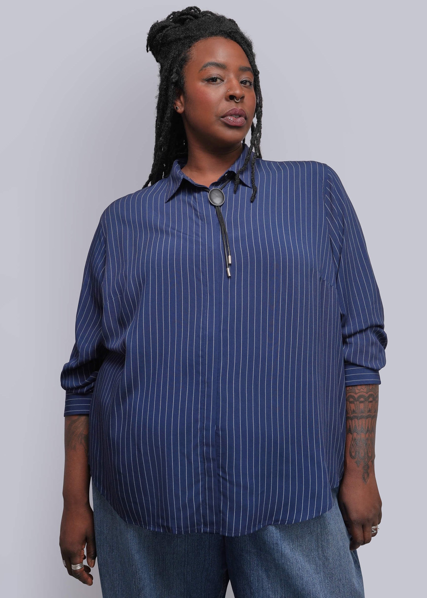 A woman modeling a blue and navy vertically striped button-down shirt with a front zipper, paired with blue jeans, against a plain light gray studio background.