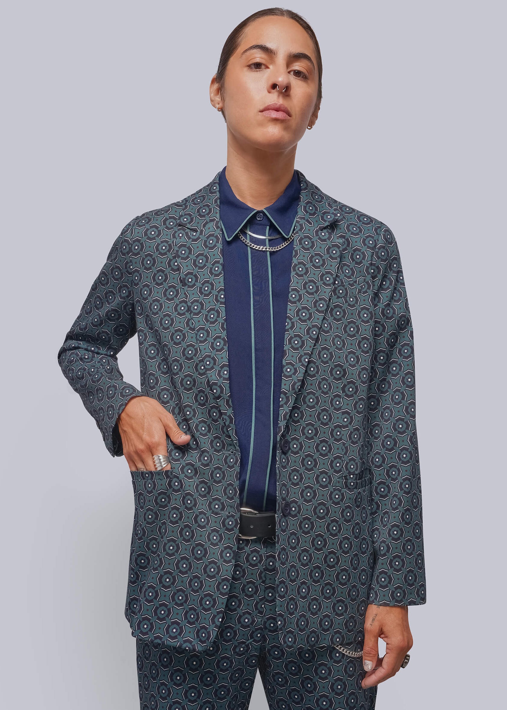 A model wearing a matching navy blue patterned blazer and trousers suit, styled with a navy shirt and a silver-toned bolo tie, against a solid gray studio background.