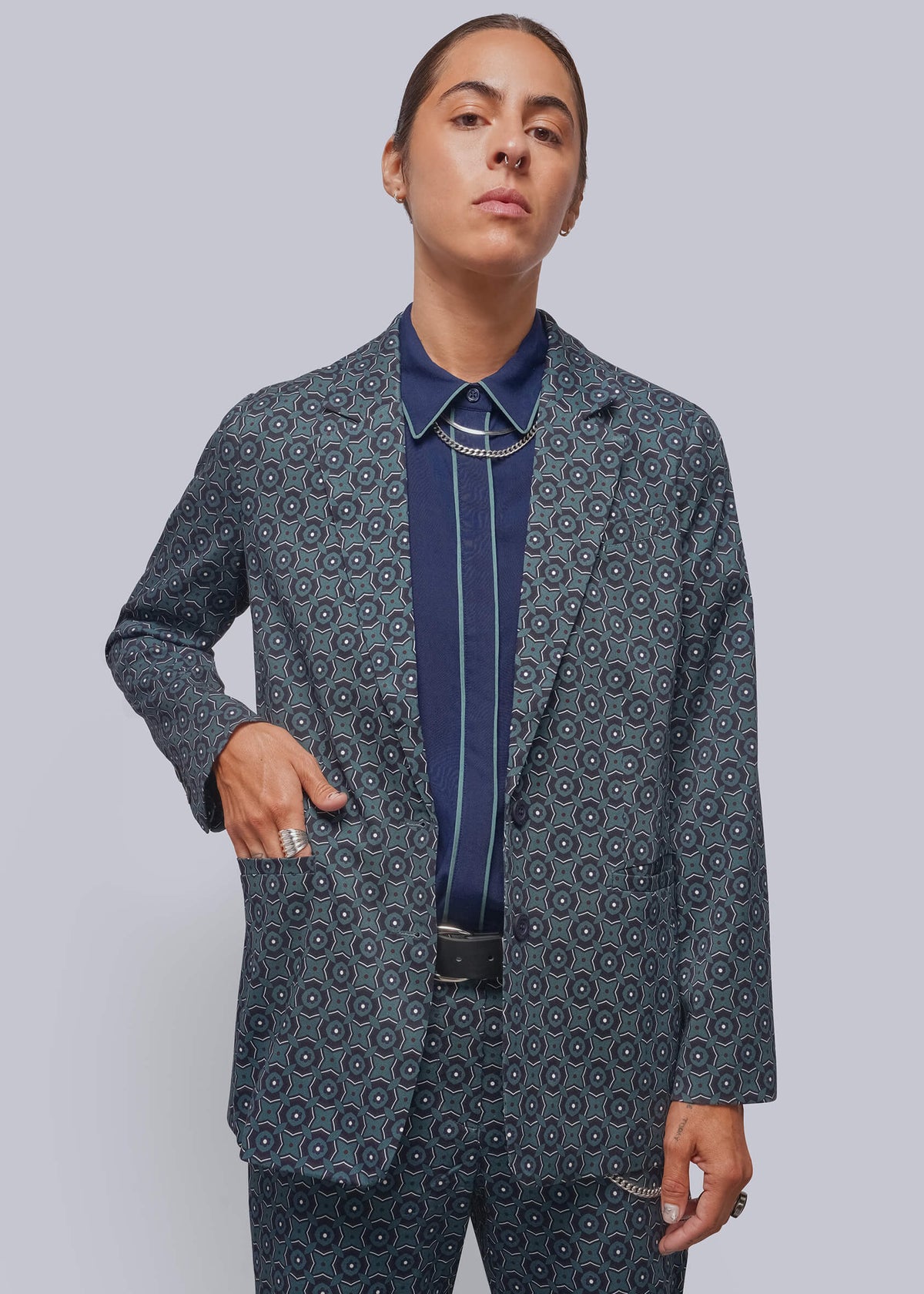 S=A model wearing a matching navy blue patterned blazer and trousers suit, styled with a navy shirt and a silver-toned bolo tie, against a solid gray studio background.