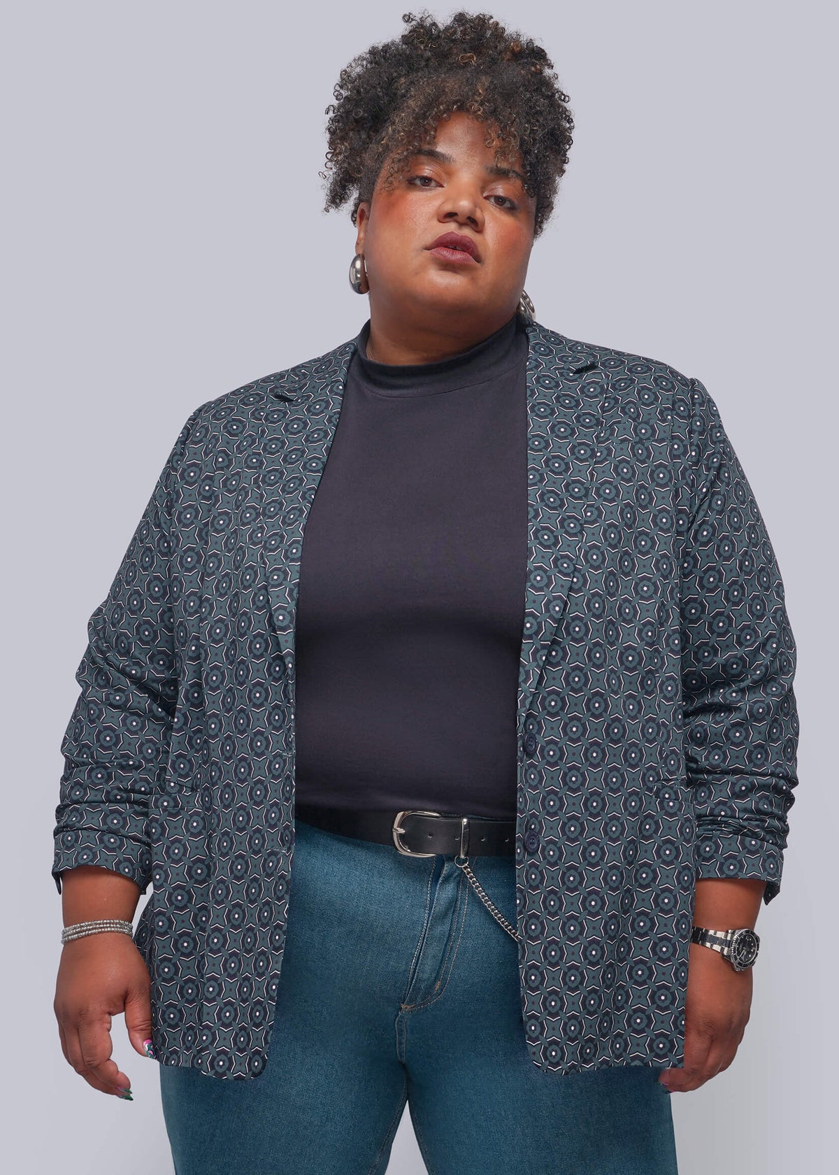 1X=A plus-size model wearing a dark gray patterned blazer over a black turtleneck and blue jeans, standing against a solid gray studio background.