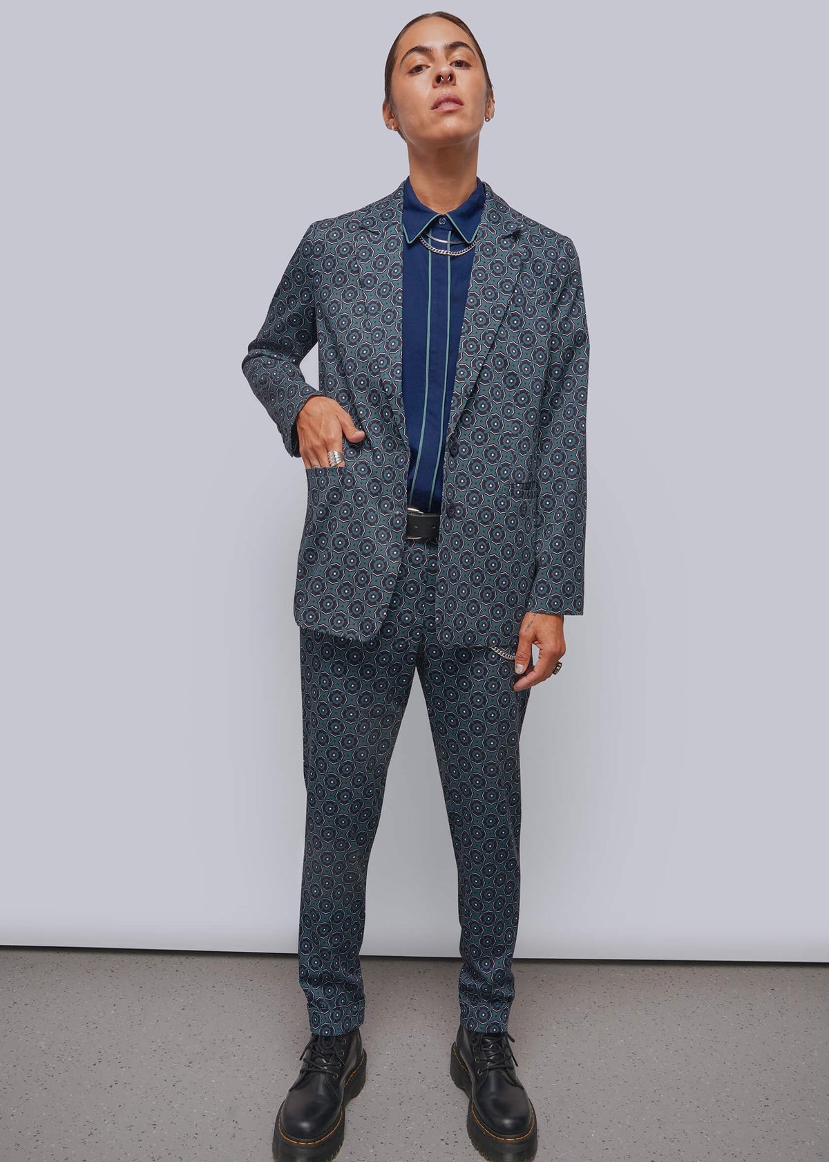S=A model stands against a light gray studio wall wearing a matching patterned blazer and trousers set in a muted blue-gray tone, layered over a dark blue collared shirt and black belt, with black leather boots.