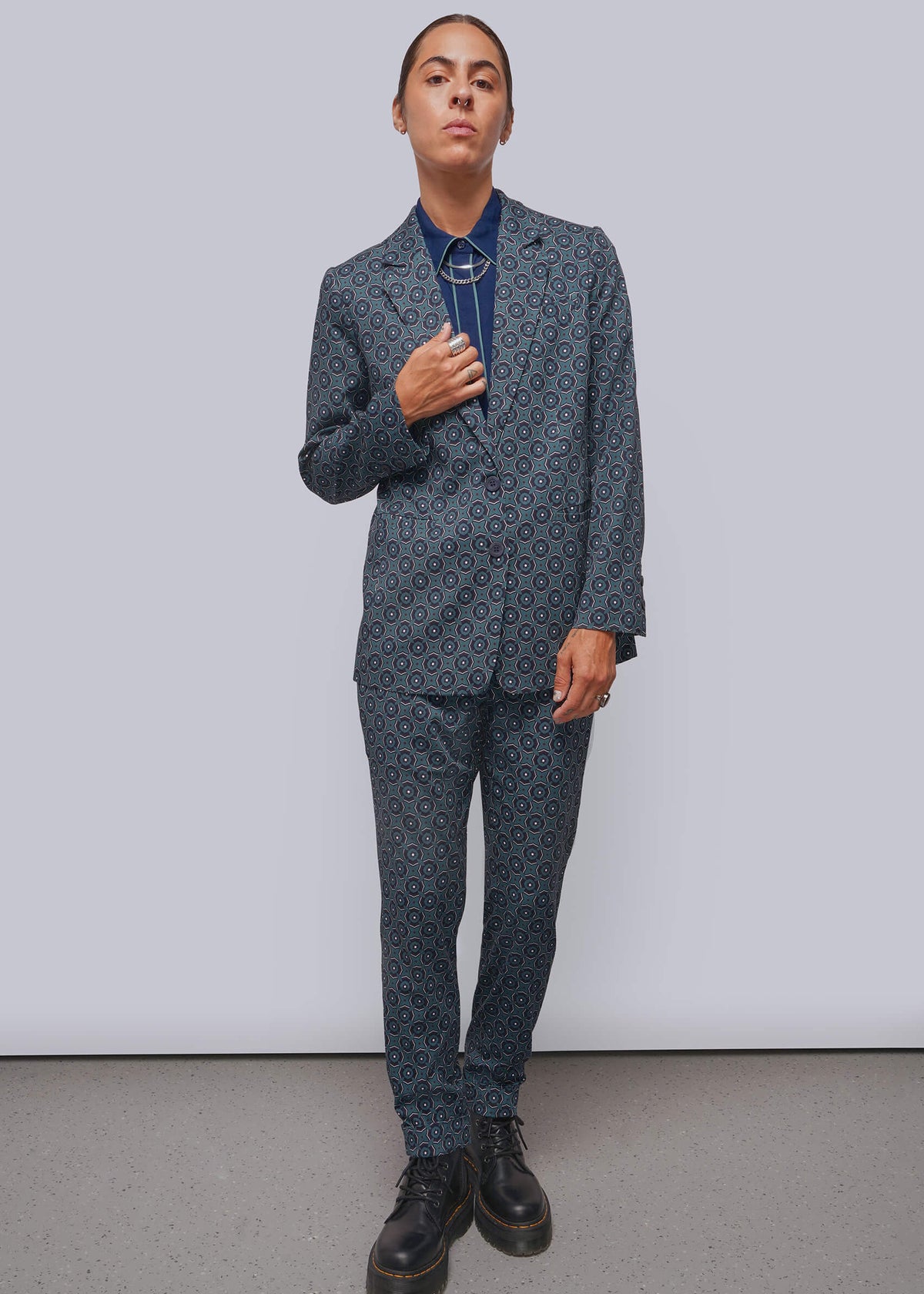 S=A model stands against a light gray studio wall wearing a matching dark blue patterned suit with a blue shirt and black boots.