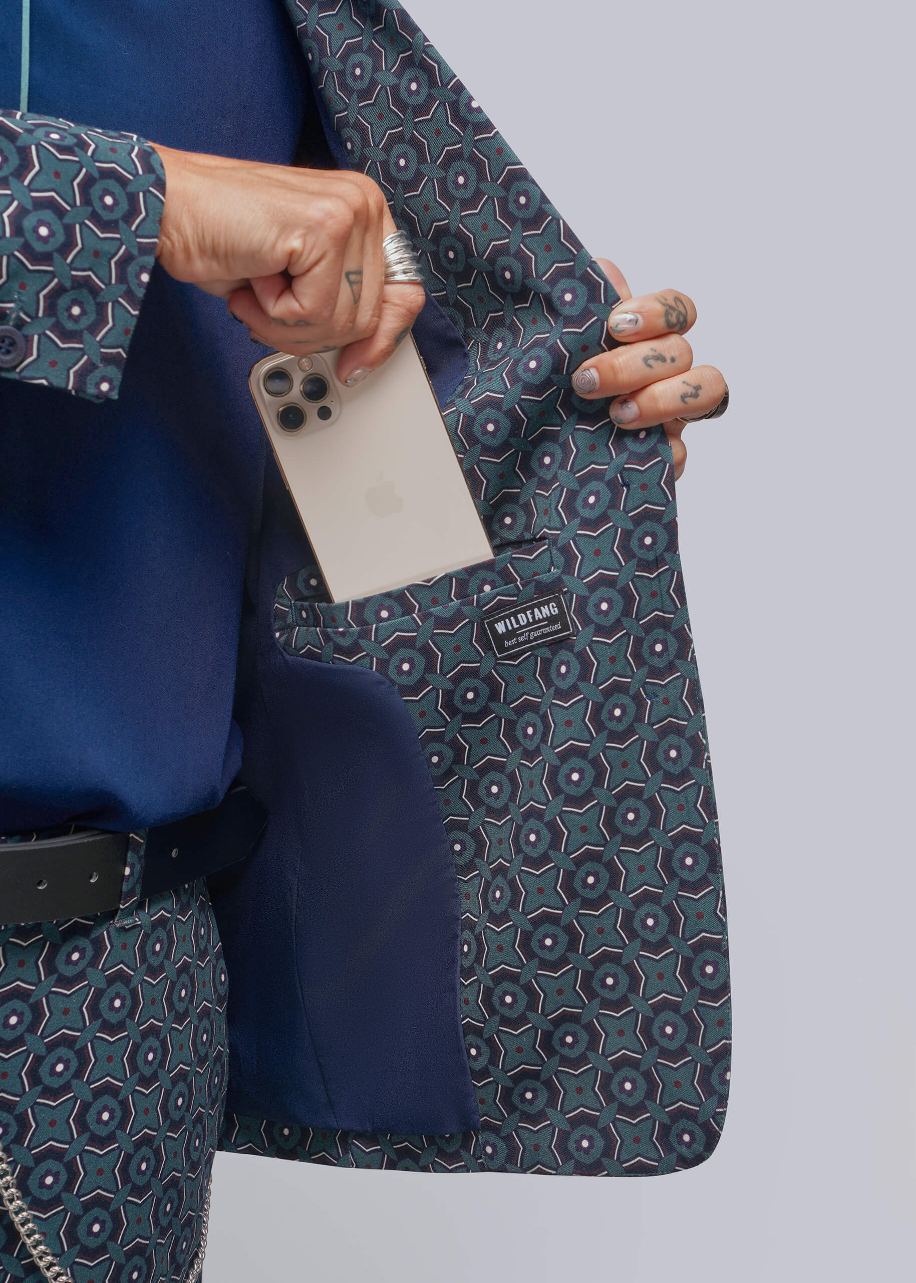 A close-up of a person wearing a blue and gray patterned blazer with a matching patterned shirt, holding a white smartphone inside the pocket to demonstrate the spacious interior pocket.