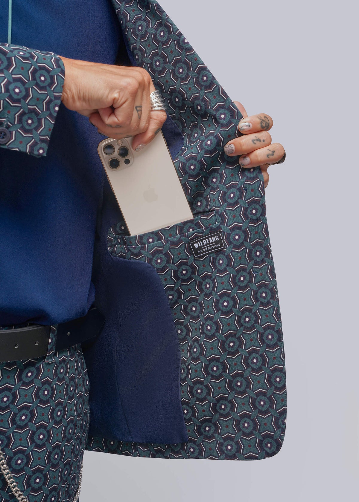 S=A close-up of a person wearing a blue and gray patterned blazer with a matching patterned shirt, holding a white smartphone inside the pocket to demonstrate the spacious interior pocket.
