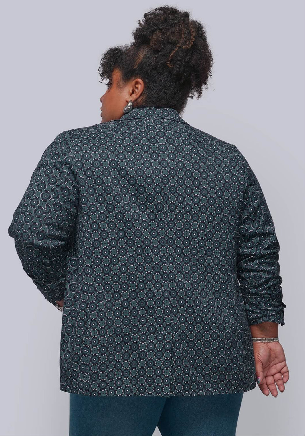 1X=A woman shown from the back wearing a dark teal button-up shirt with a repeating circular medallion pattern, paired with blue jeans, against a solid light gray background.