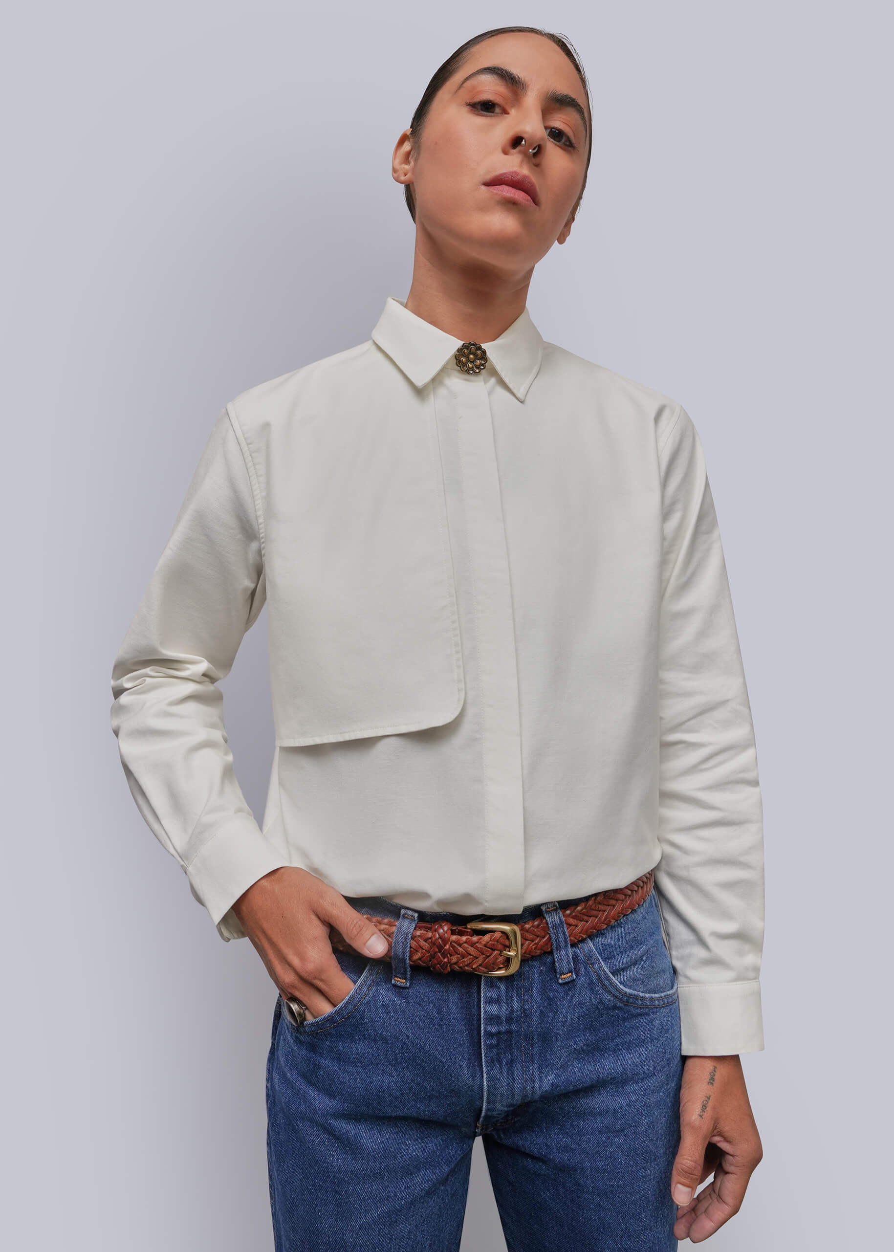 A model wearing a white long-sleeve button-down shirt with a chest pocket and a decorative button at the collar, paired with blue jeans and a brown woven belt, against a solid gray background.