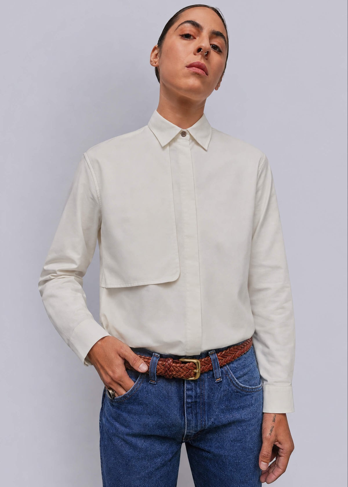 S=A model wearing a white long-sleeve button-down shirt, paired with blue jeans and a brown woven belt, against a solid gray background.