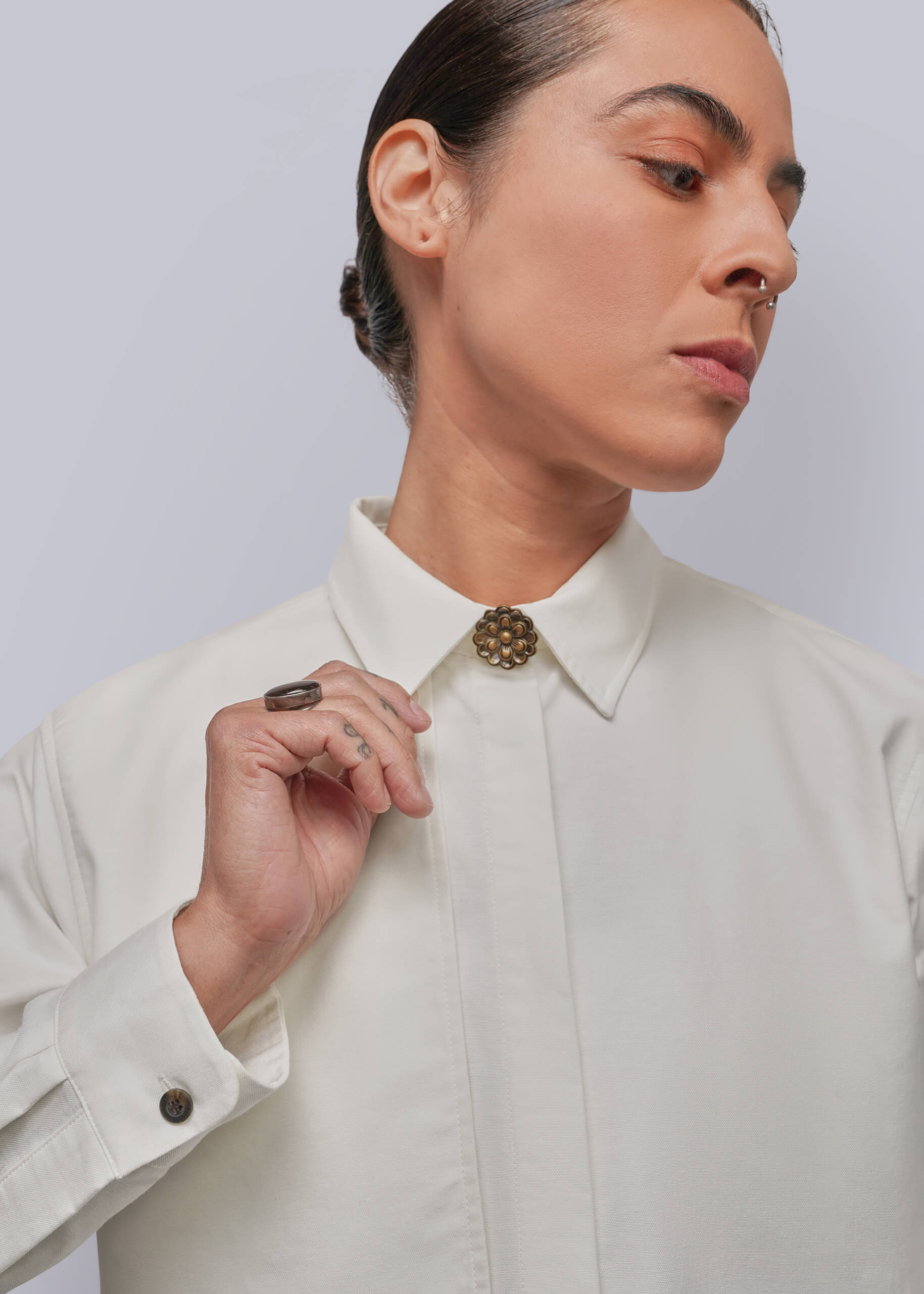 S=A close-up of a model wearing a white long-sleeve button-down shirt with a decorative metallic circular button cover at the collar, set against a plain light gray background.