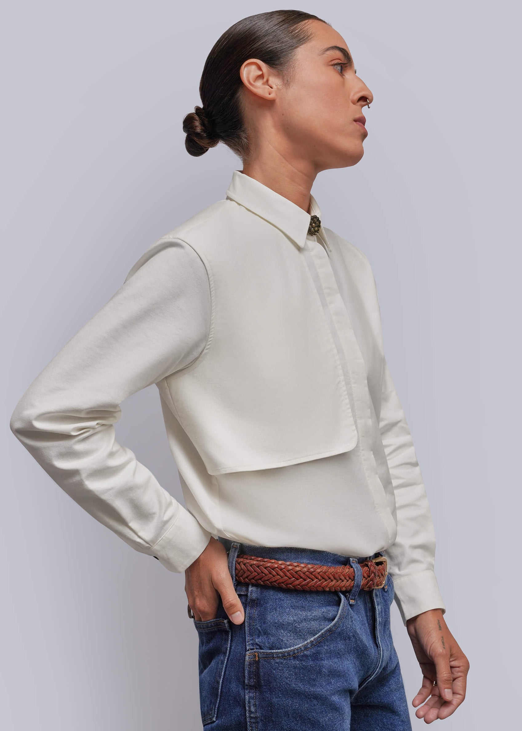 S=A model shown in profile wearing a cream-colored long-sleeve button-up shirt with a storm flap chest panel, paired with blue jeans and a brown braided belt, against a solid light gray background.