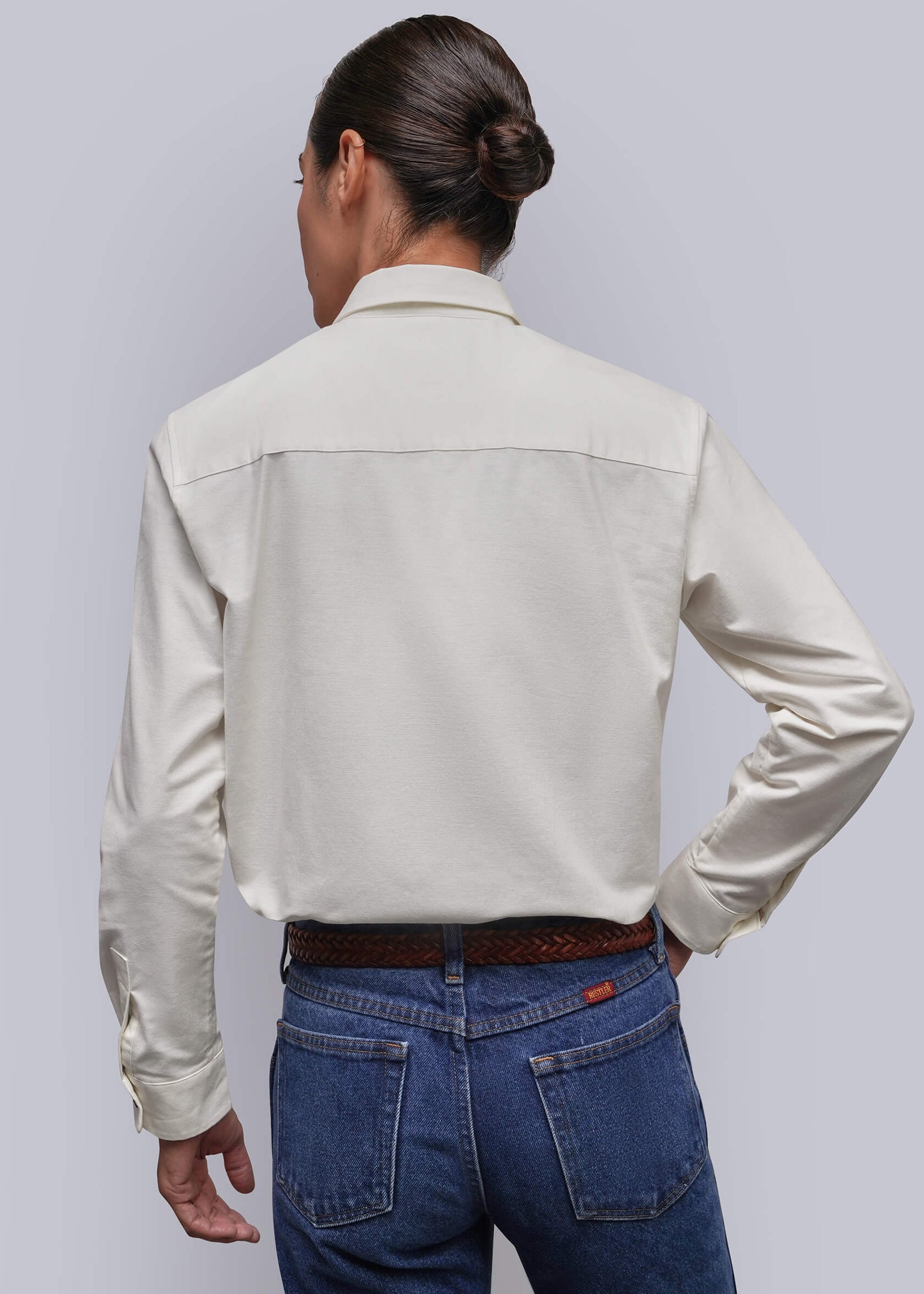 S=A person viewed from the back wearing a light cream-colored long-sleeve button-down shirt and blue jeans, standing against a solid light gray background.