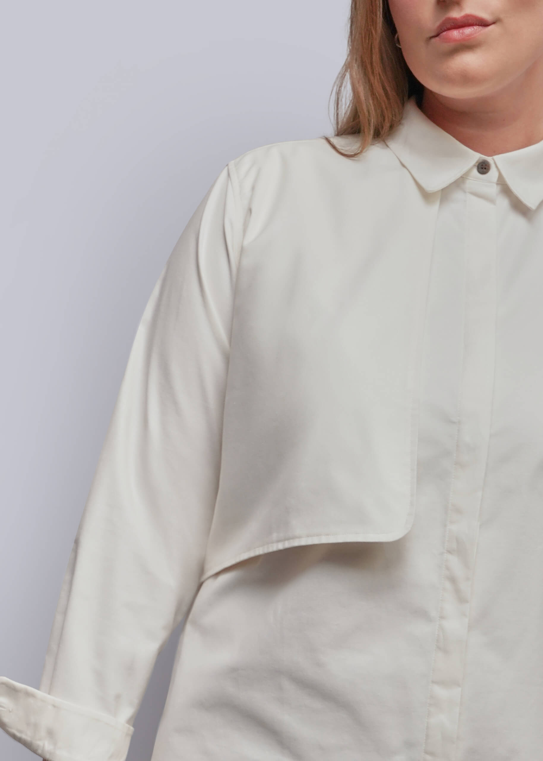1X=A close-up of a model wearing a light cream or off-white long-sleeve button-up shirt with a collar and a unique asymmetrical draped pocket detail on the chest, set against a plain light gray background.