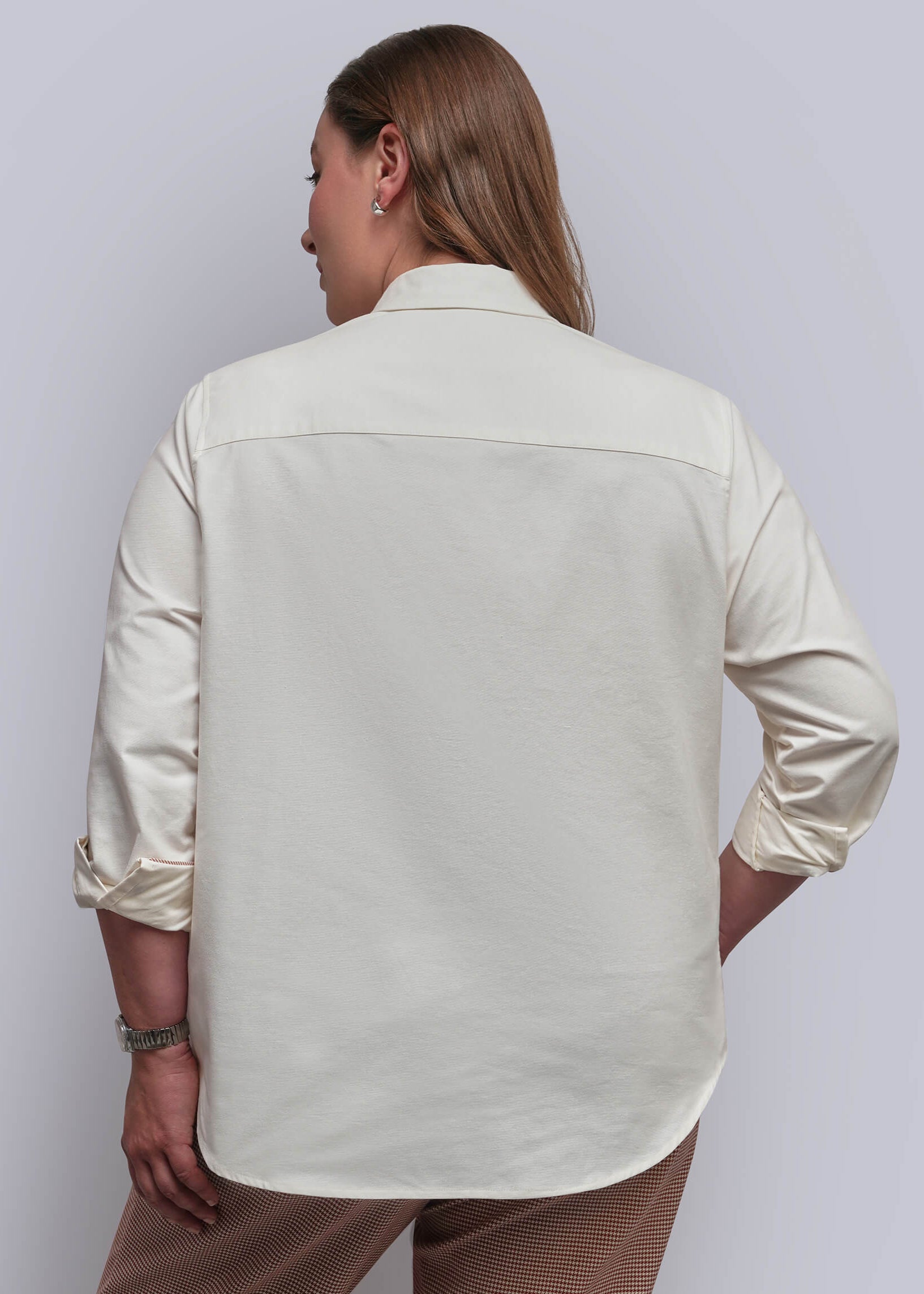 1X=A woman shown from the back wearing a light cream-colored long-sleeve button-down shirt and houndstooth pants, set against a plain light gray studio background.